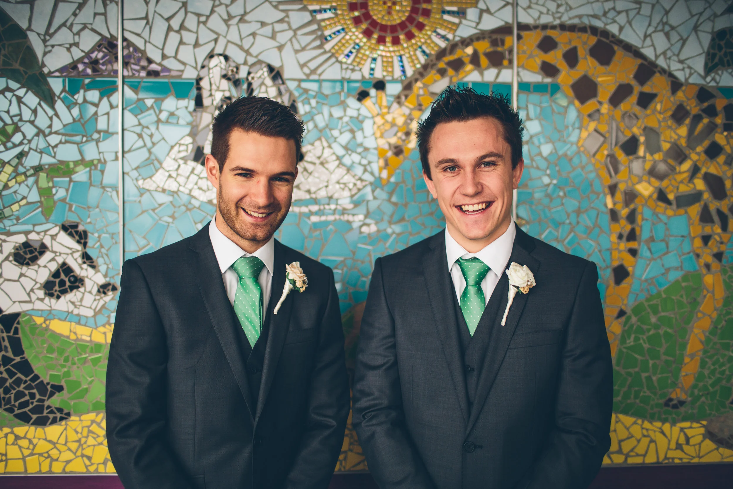Two young men in suits with green ties and boutonnières, smiling, standing in front of a colorful mosaic mural.