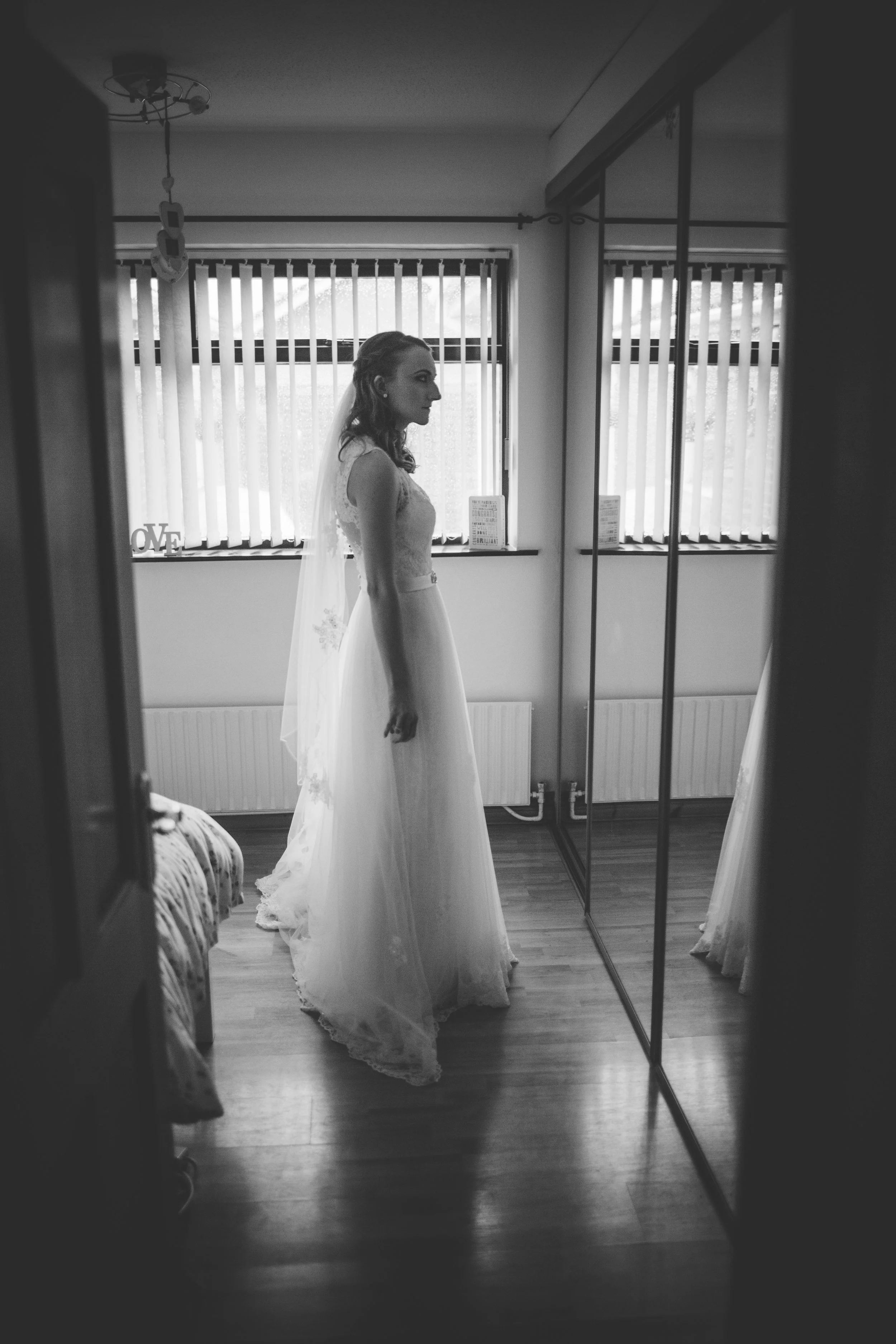 Black-and-white photo of a woman in a wedding dress standing in front of a mirror, with windows and vertical blinds in the background.