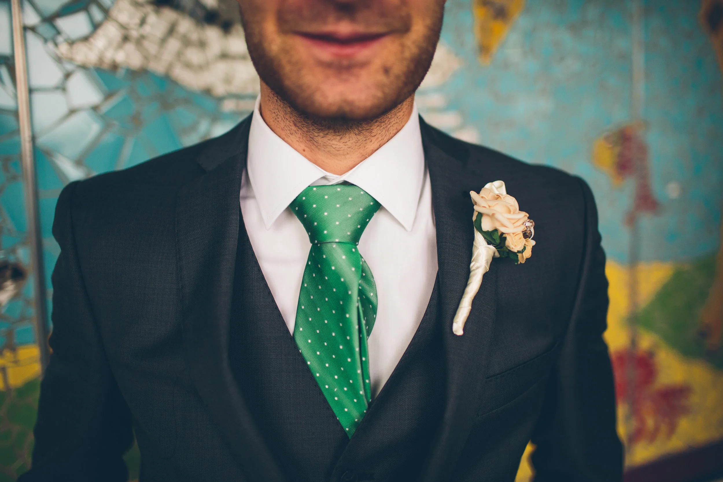 A man in a dark suit with a white shirt and green polka dot tie, wearing a boutonniere, standing in front of a colorful wall.