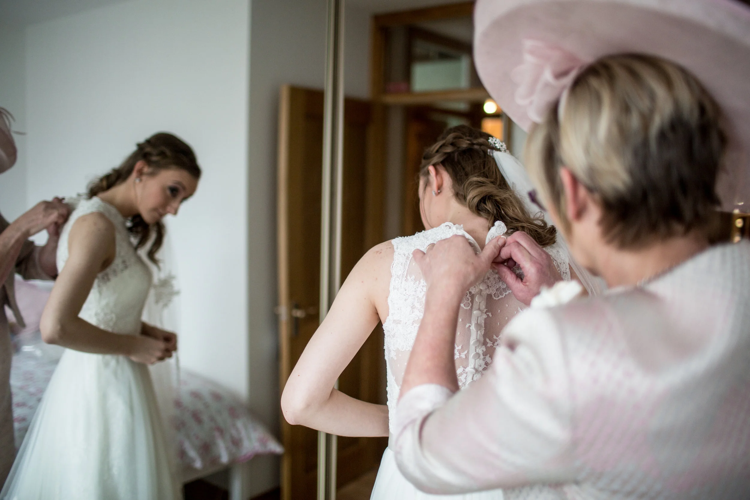 A bride in a lace wedding dress is being helped by an older woman with her dress at a mirror, with another woman helping in the background.