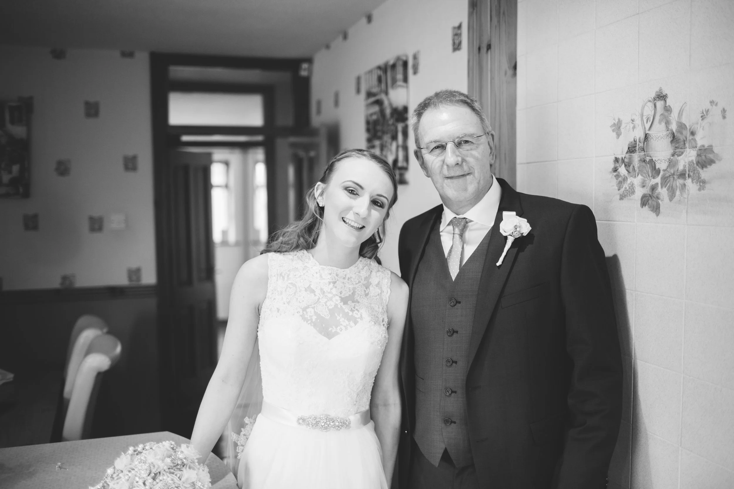 A bride and an older man, possibly her father, standing together indoors. The woman is wearing a lace wedding dress and smiling. The man is dressed in a suit with a boutonniere, also smiling.