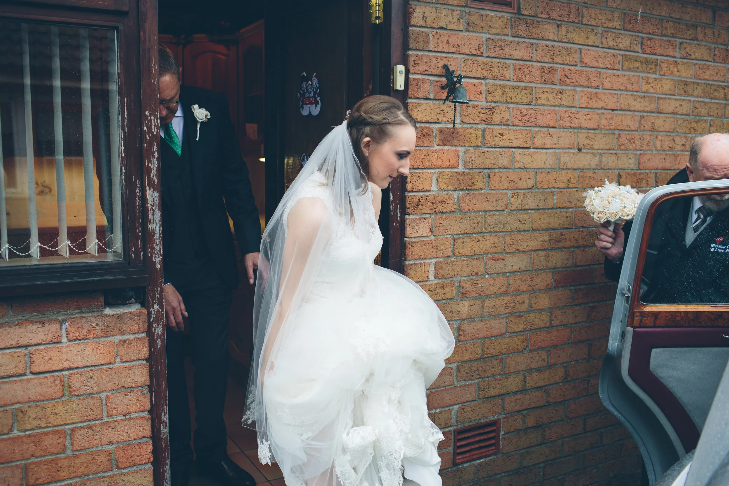 Bride in a white wedding gown and veil stepping out of a house with a brick wall, while an elderly man holds a bouquet of white roses nearby.