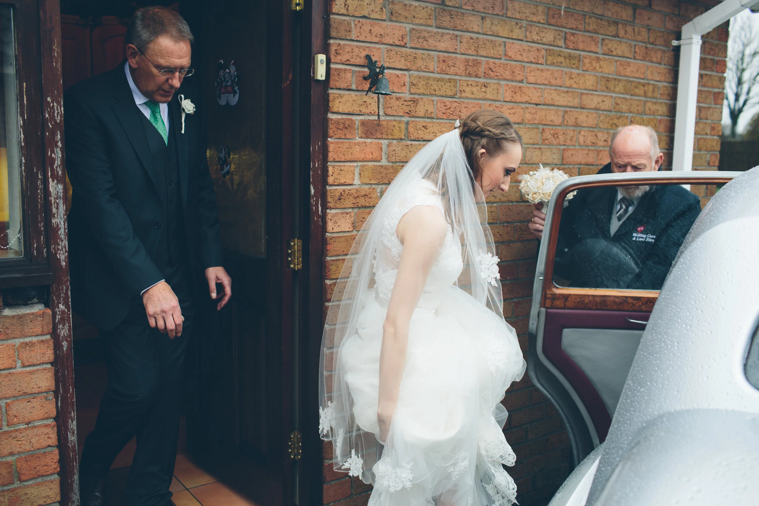 Bride in a white wedding gown and veil getting into a car with assistance from an older man. Other man holding a bouquet of flowers standing behind the car, outside a brick house.