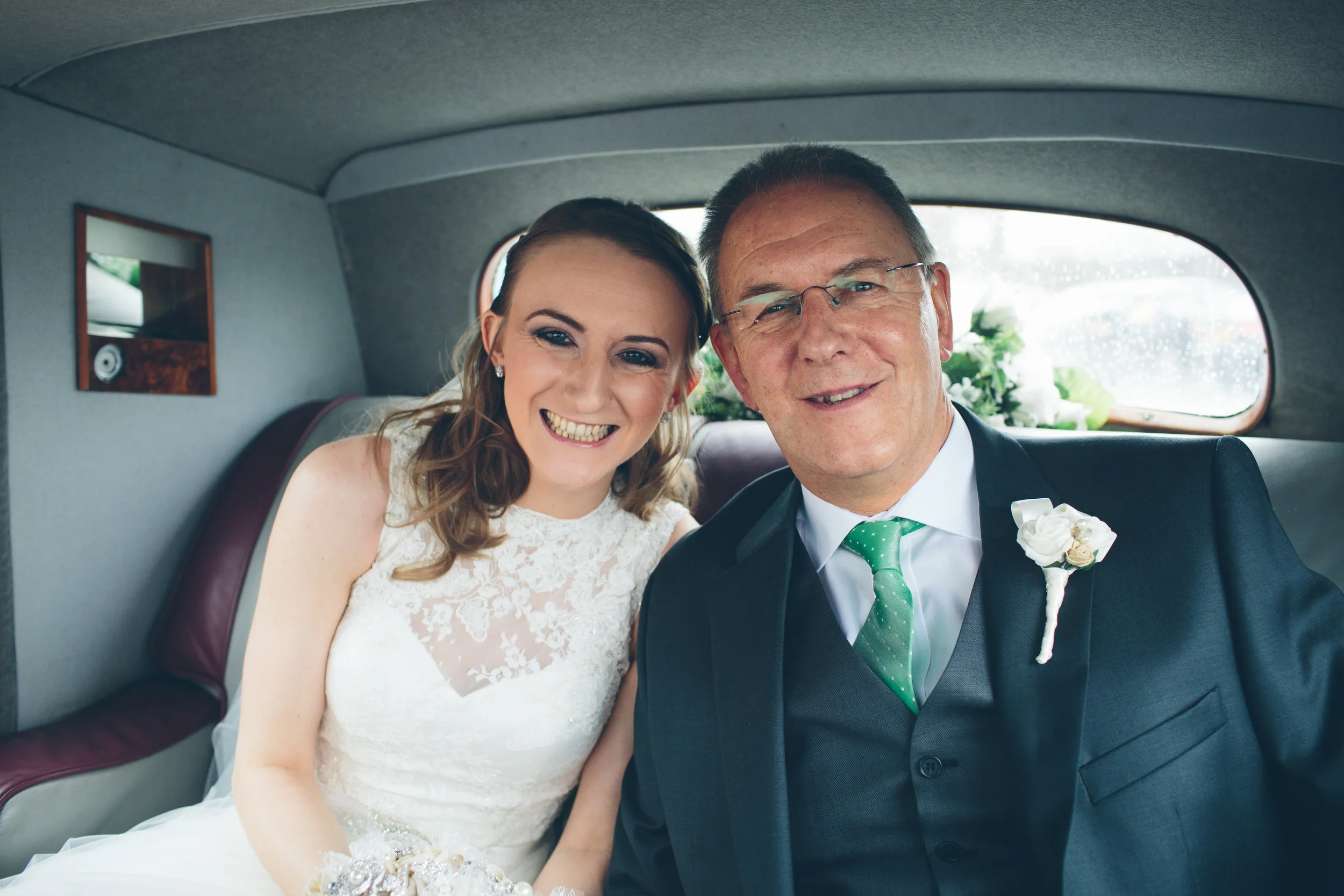 A bride and an older man, possibly her father, sitting inside a car dressed for a wedding, both smiling.