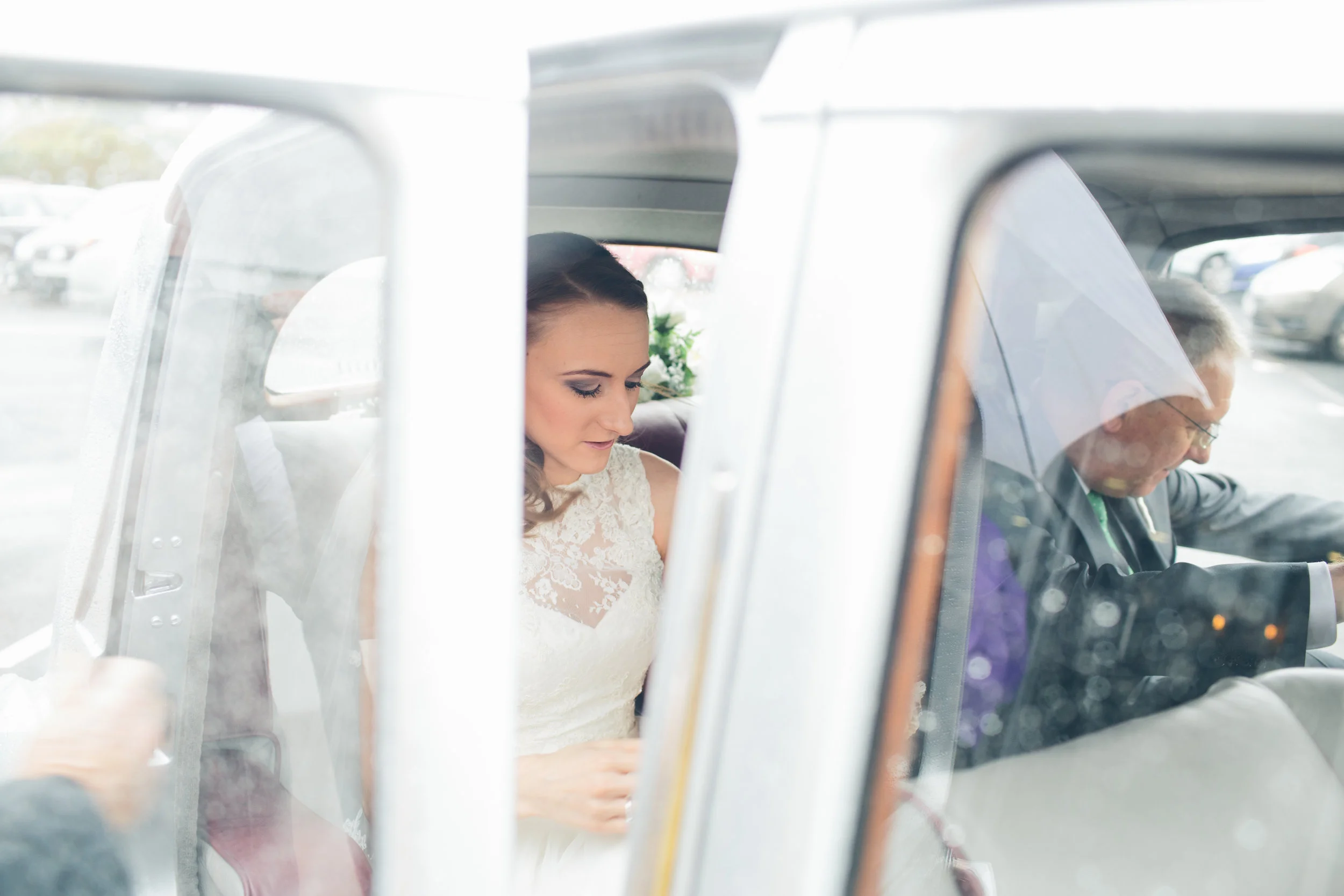 Bride in white dress sitting inside a car, with a man driving, seen through a car window.