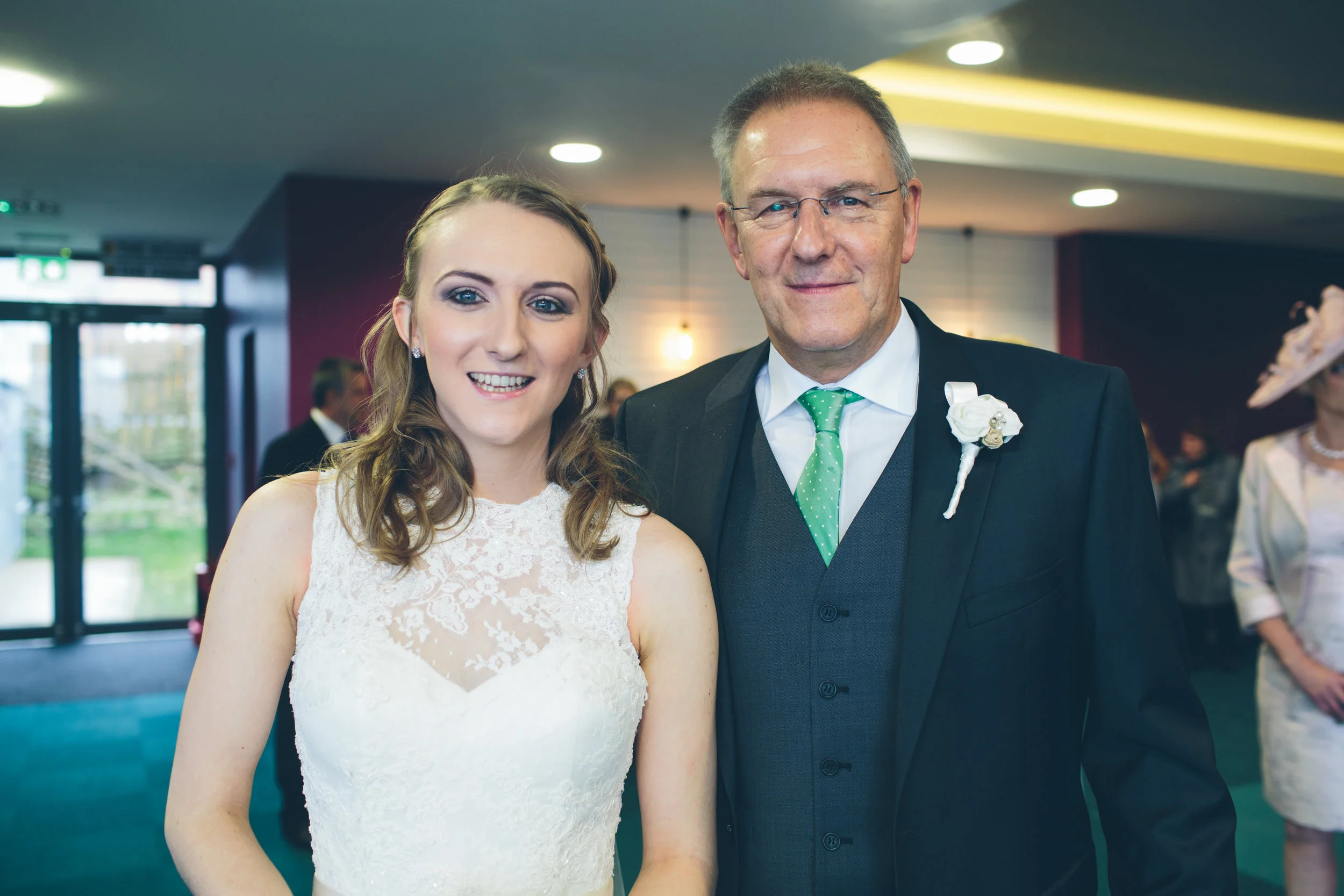 A young woman in a white wedding dress standing next to an older man in a black suit with a green tie and white boutonniere at a wedding reception.