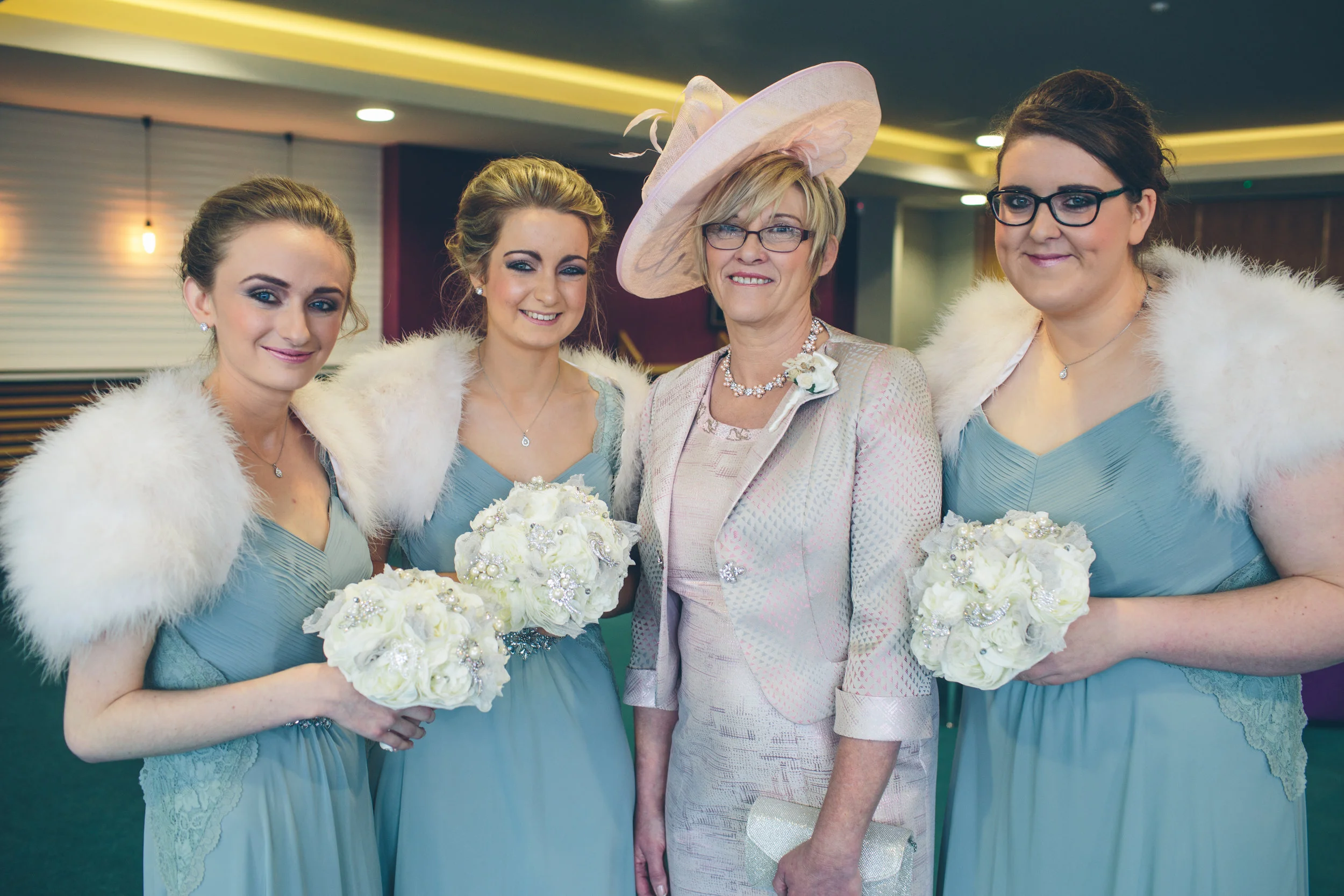 Four women dressed in formal attire, three holding bouquets, standing together indoors, at a wedding or special event.