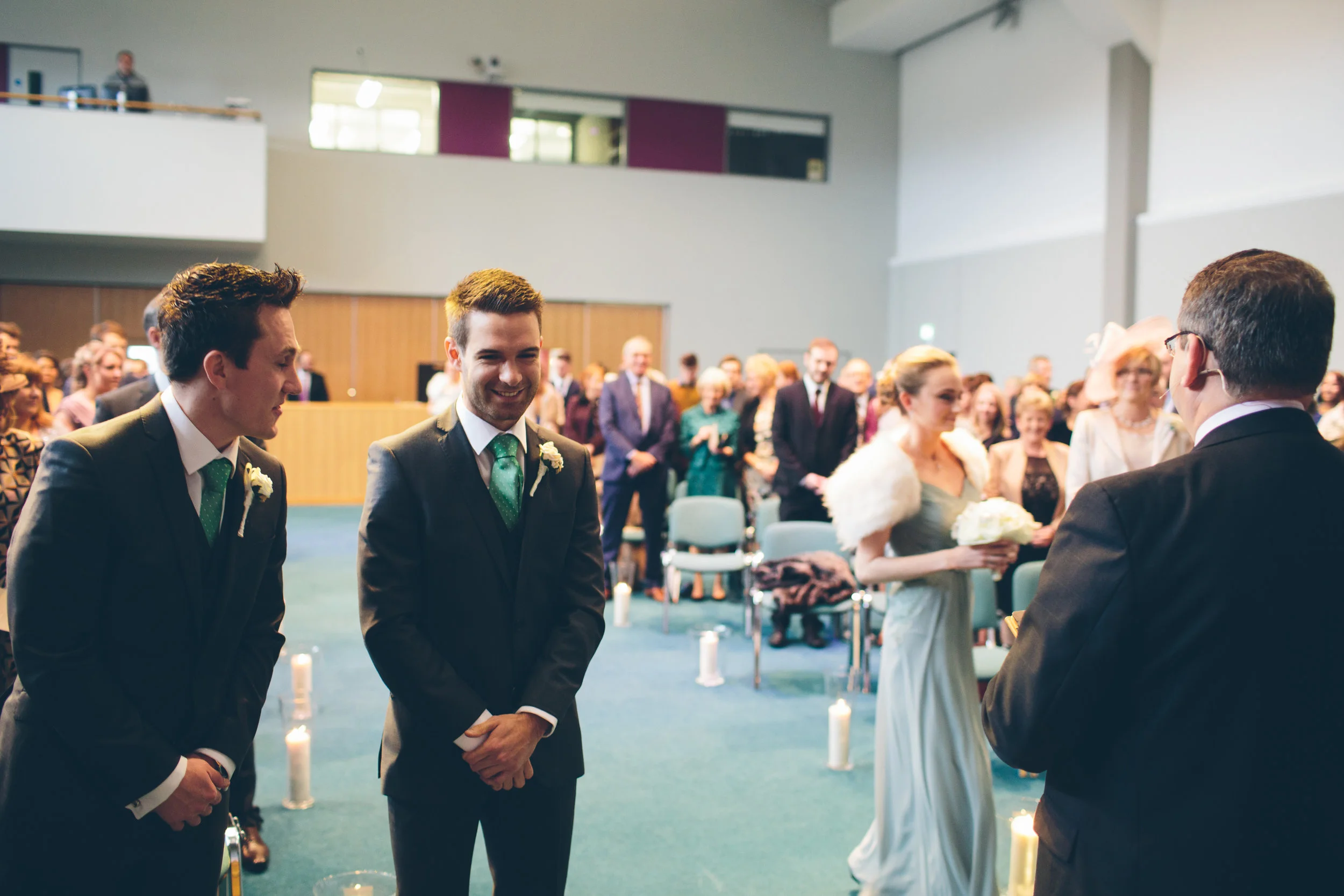 A wedding ceremony with two grooms in suits and a bride in a light-colored dress holding a bouquet, facing a man in a tuxedo, while guests stand and watch in a decorated indoor space.