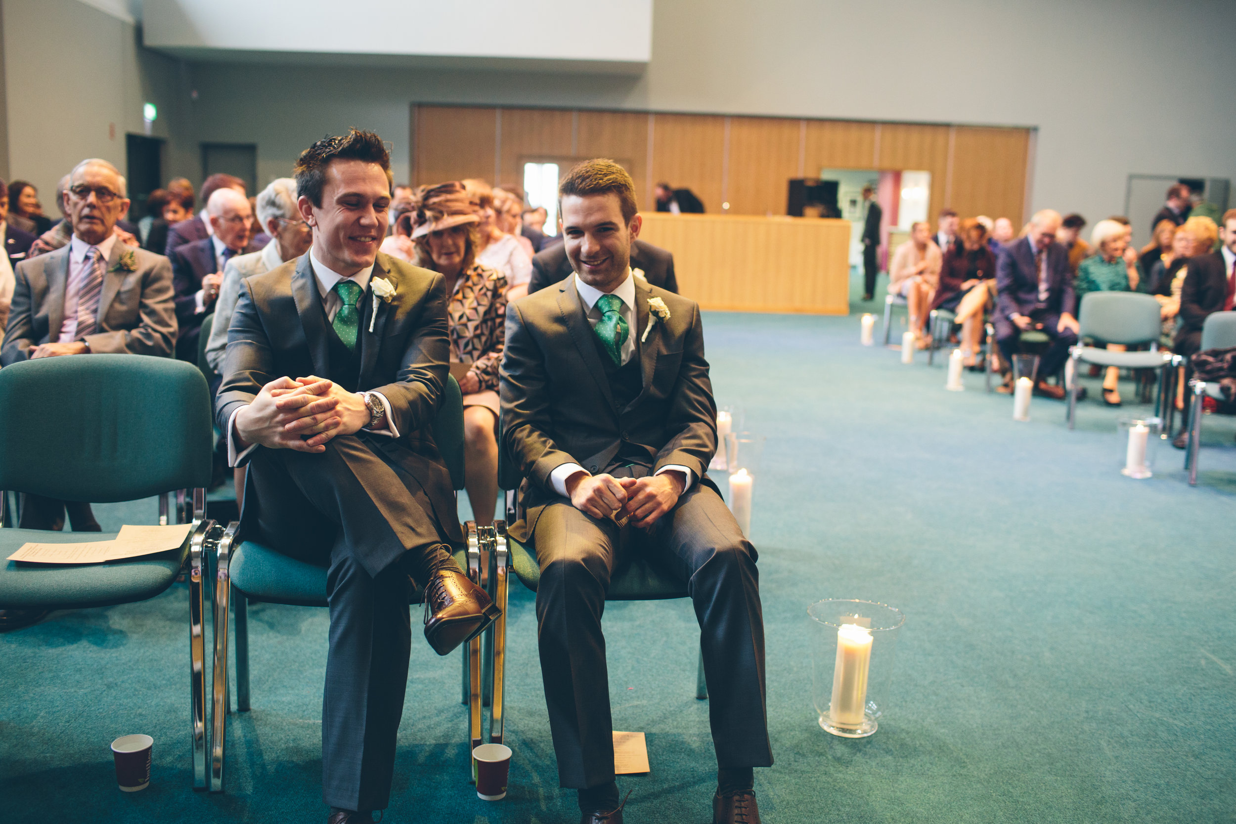 Two men in suits sitting in the front row at a wedding ceremony, smiling and talking to each other, with guests seated behind them and candles on the floor.