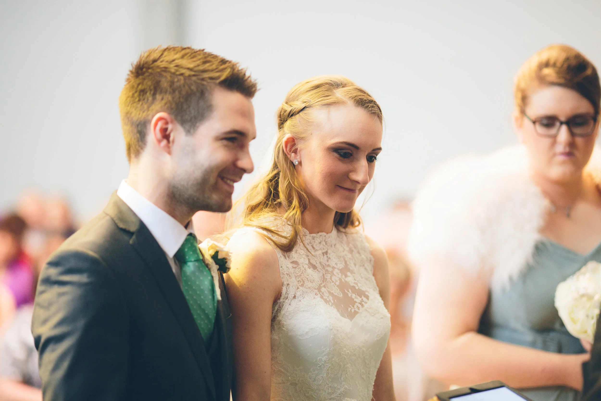 A bride and groom standing together during their wedding ceremony, smiling and looking thoughtful, with a woman on the right holding a bouquet in the background.
