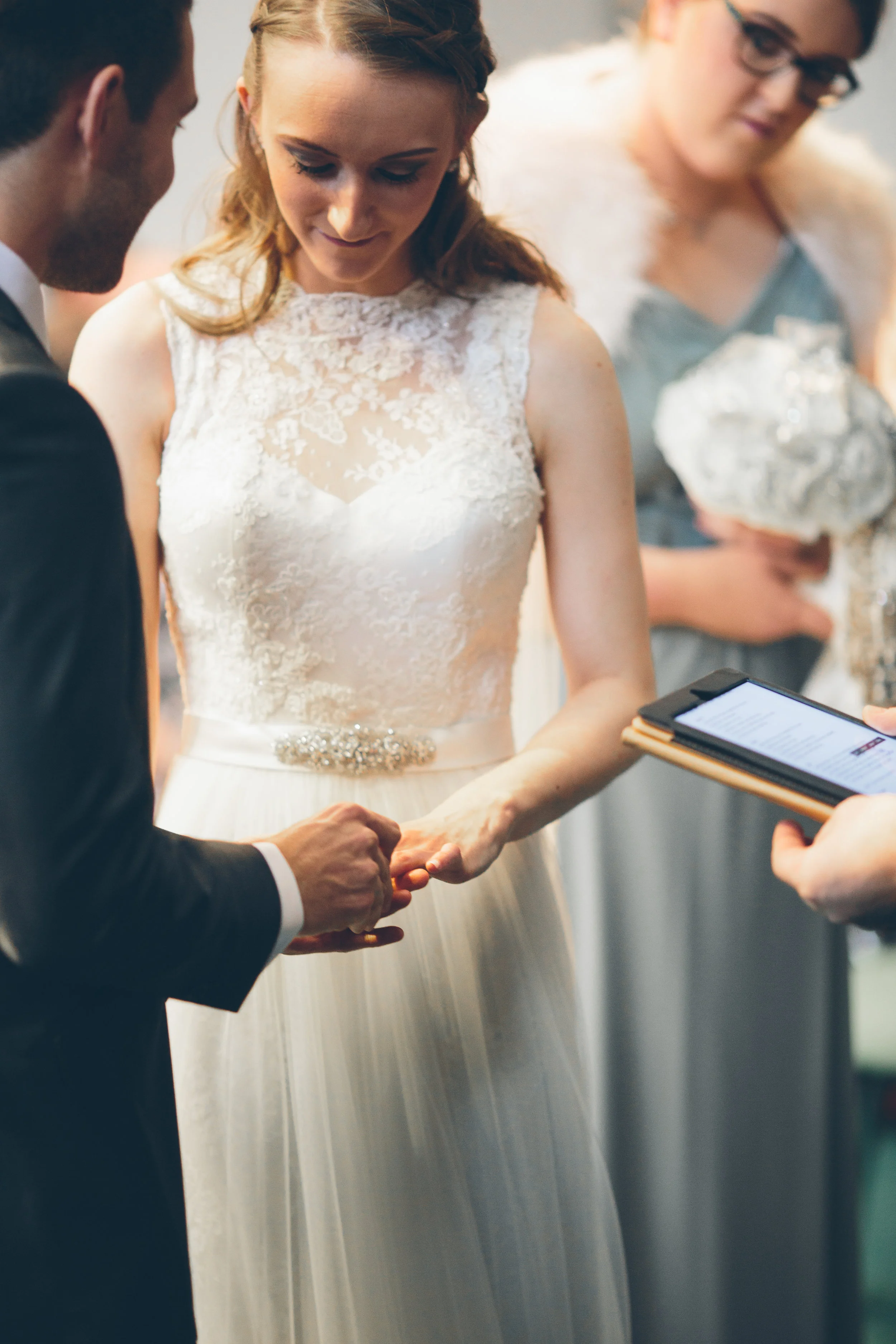 A wedding ceremony where a bride and groom are exchanging rings, with a witness holding a tablet nearby.