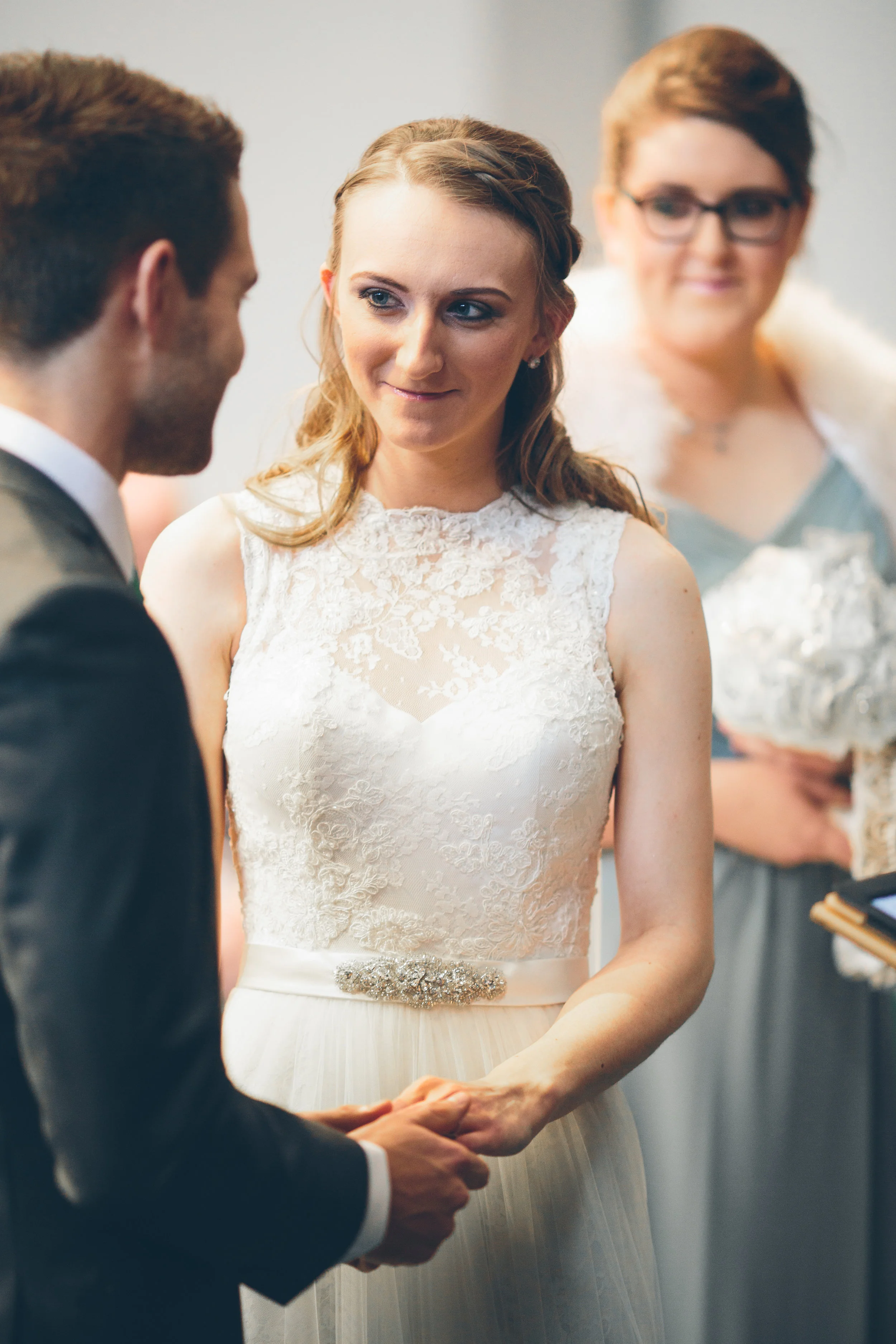 A bride in a lace wedding dress holding hands with a groom in a suit during a wedding ceremony, with a woman in the background holding a bouquet.