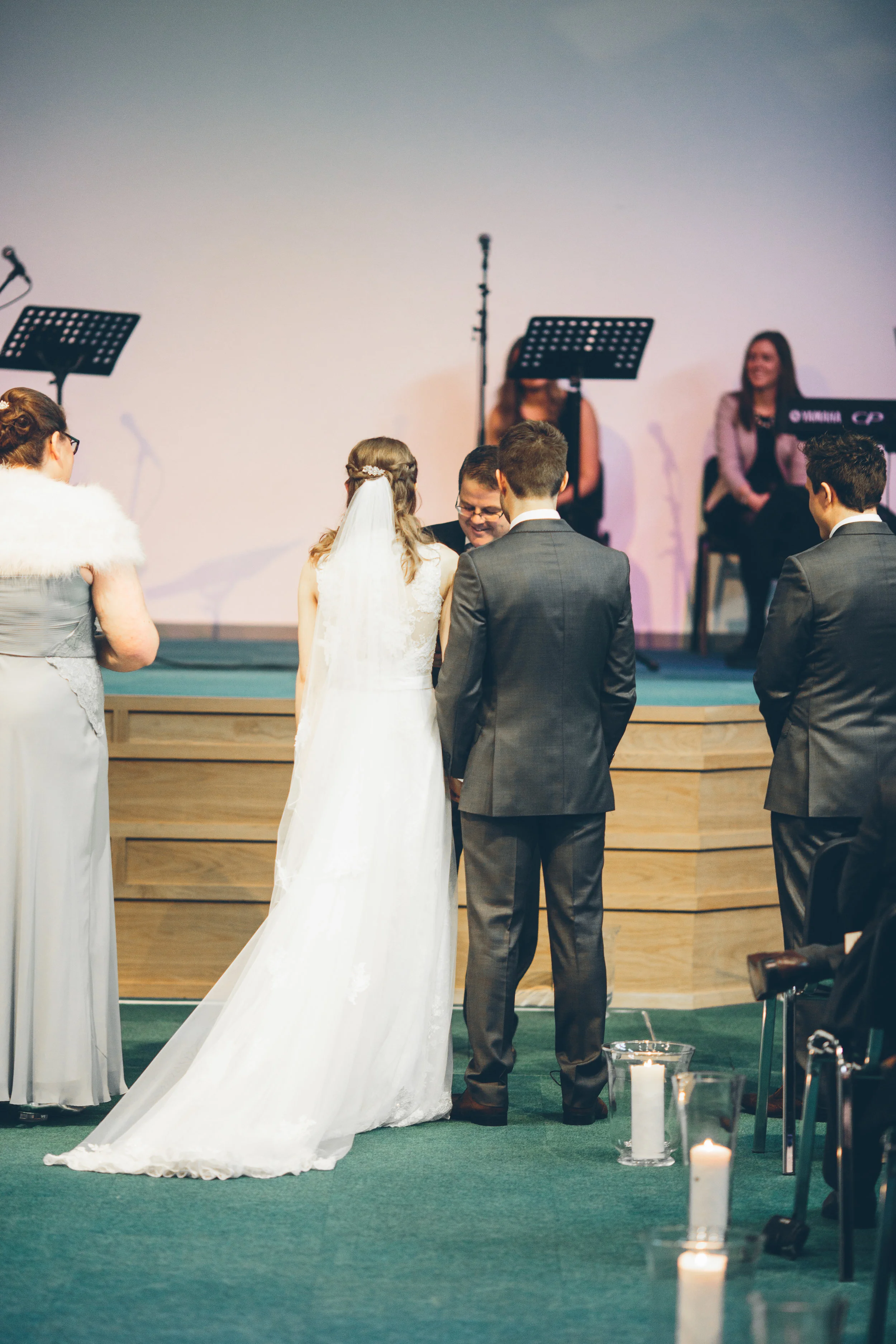 A wedding ceremony with a bride and groom exchanging vows, surrounded by friends and a seated officiant, with musicians in the background.