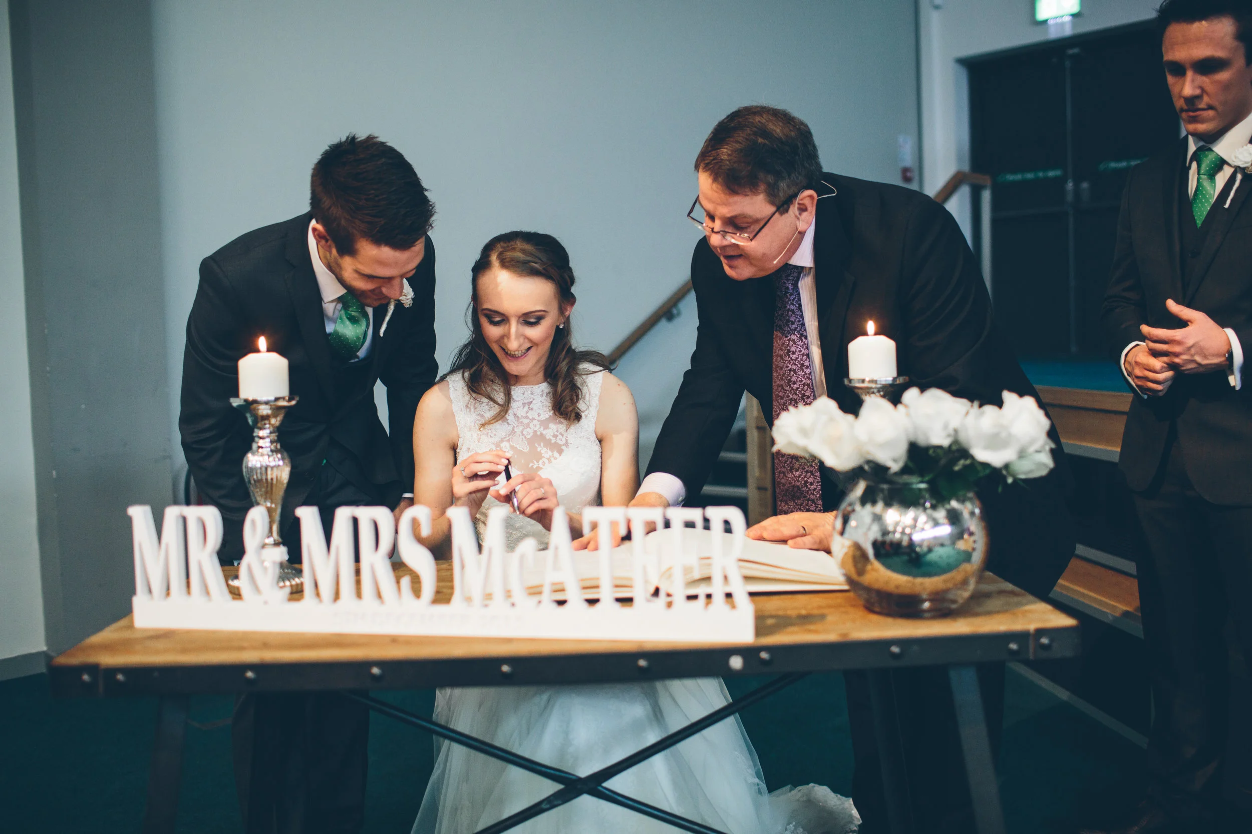 Wedding reception with bride and groom signing a marriage certificate, surrounded by two men in suits, with candles and a sign that reads 'Mr. & Mrs. McAtter' on the table.