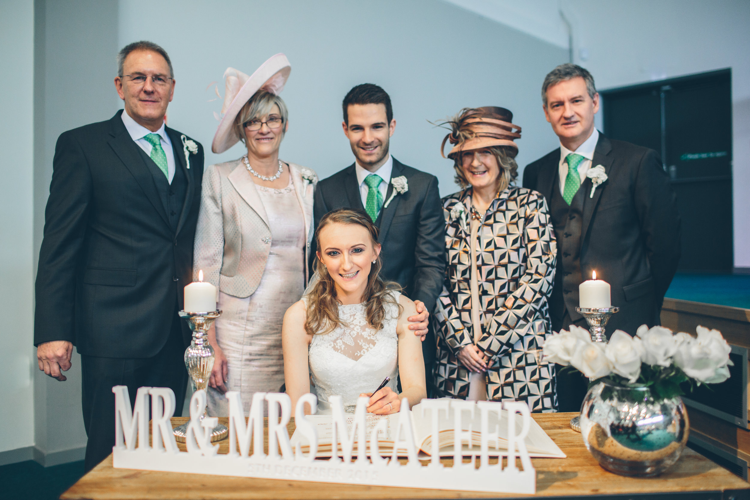 A newly married woman in a white wedding dress sits at a table with a pen, signing a document, as her family and friends stand behind her, smiling. The table is decorated with white roses and candles. A sign on the table reads "MR & MRS MAXTER".