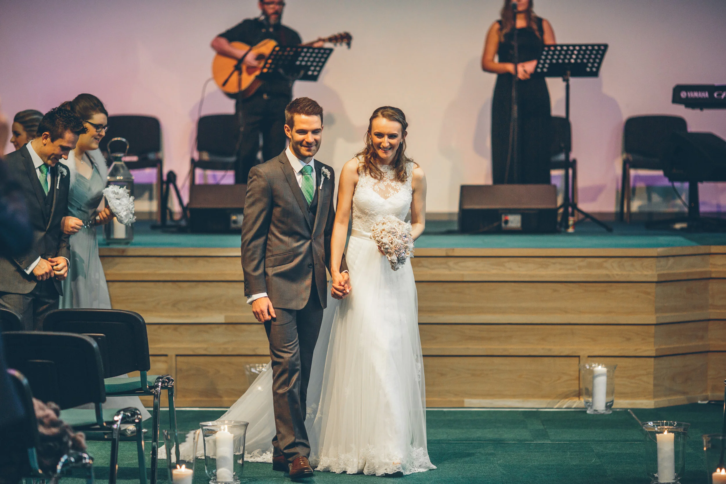 A newlywed couple is holding hands and walking down the aisle in a church during their wedding ceremony, with a small group of guests on the side and a band playing in the background.