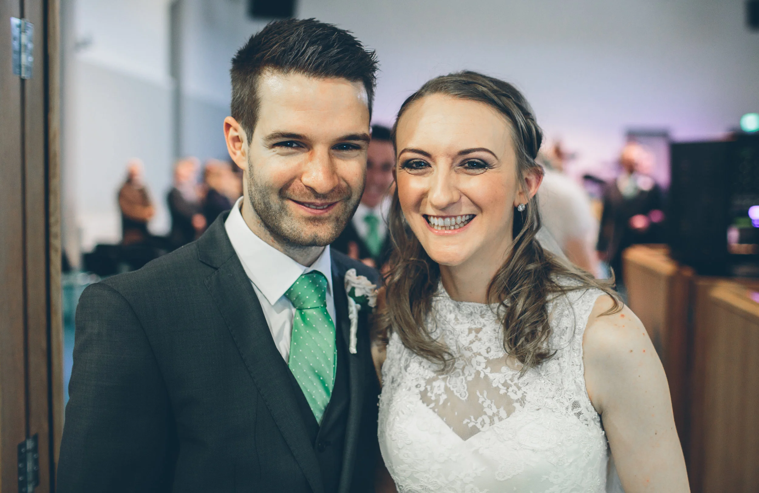 A happy couple at a wedding reception, smiling and standing close together in formal attire with blurred background of guests.