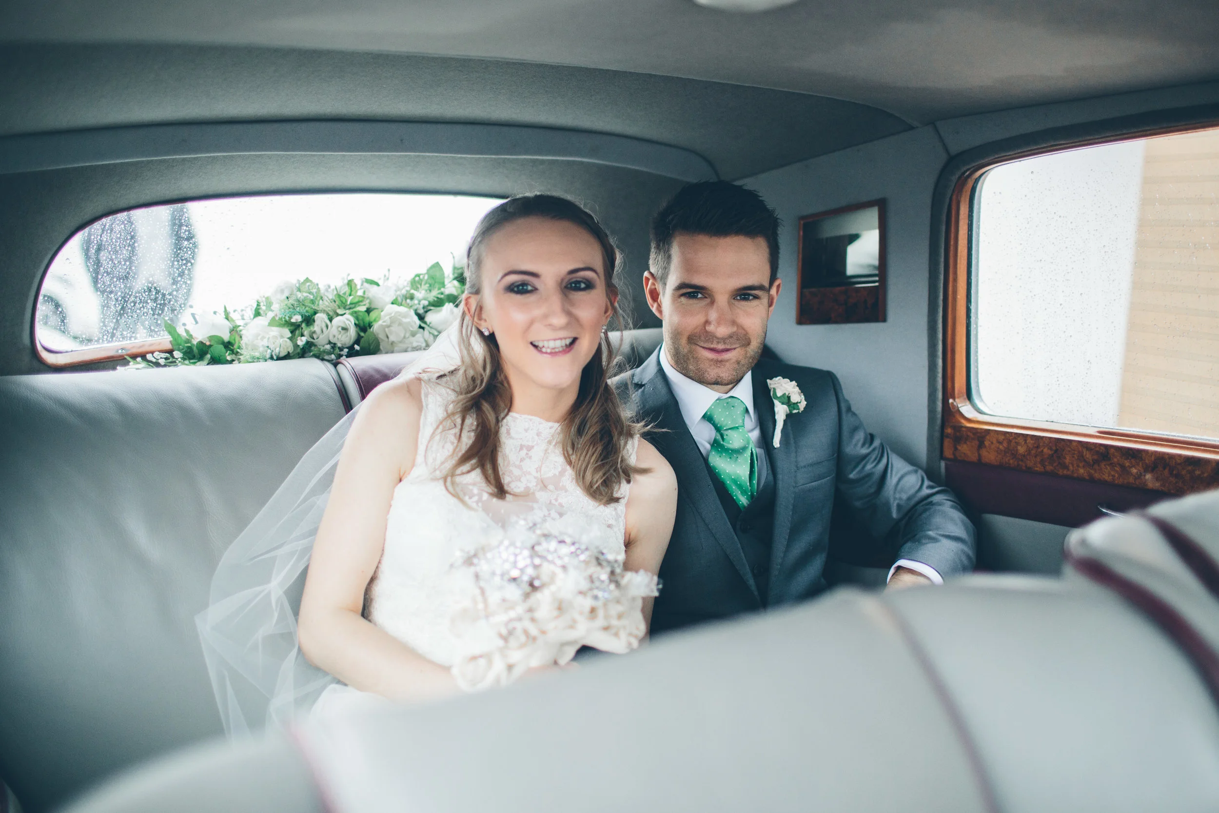 Bride and groom sitting inside a vintage car, smiling, with flowers in the background