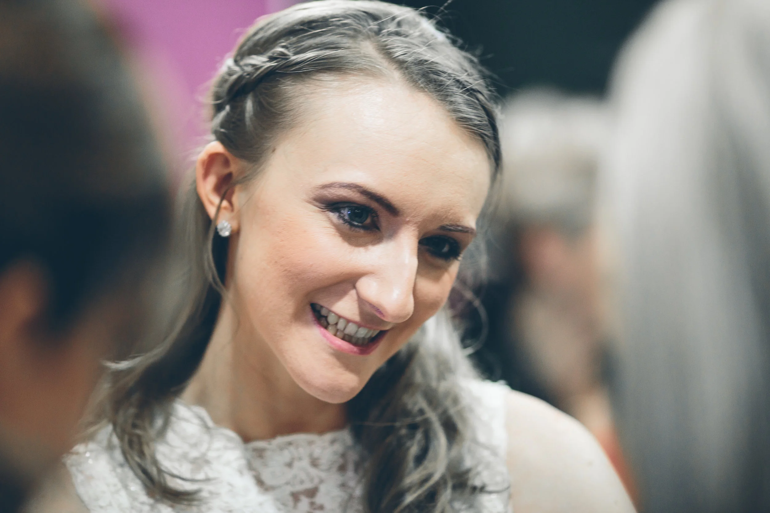 Close-up of a smiling woman with a braid hairstyle, wearing jewelry and a white lace top, in a social setting.