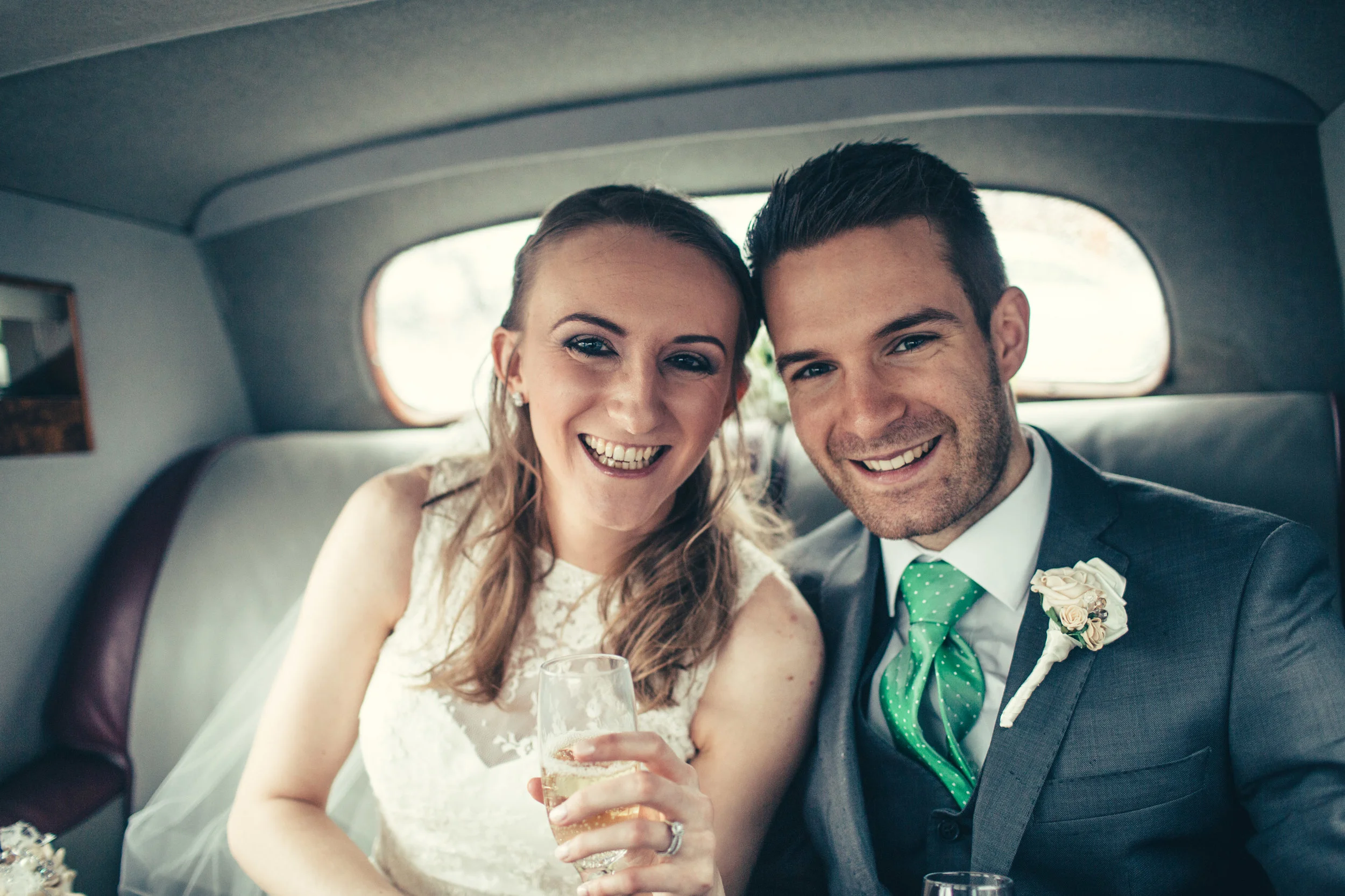 A smiling bride and groom sitting inside a vehicle, celebrating their wedding with glasses of champagne.