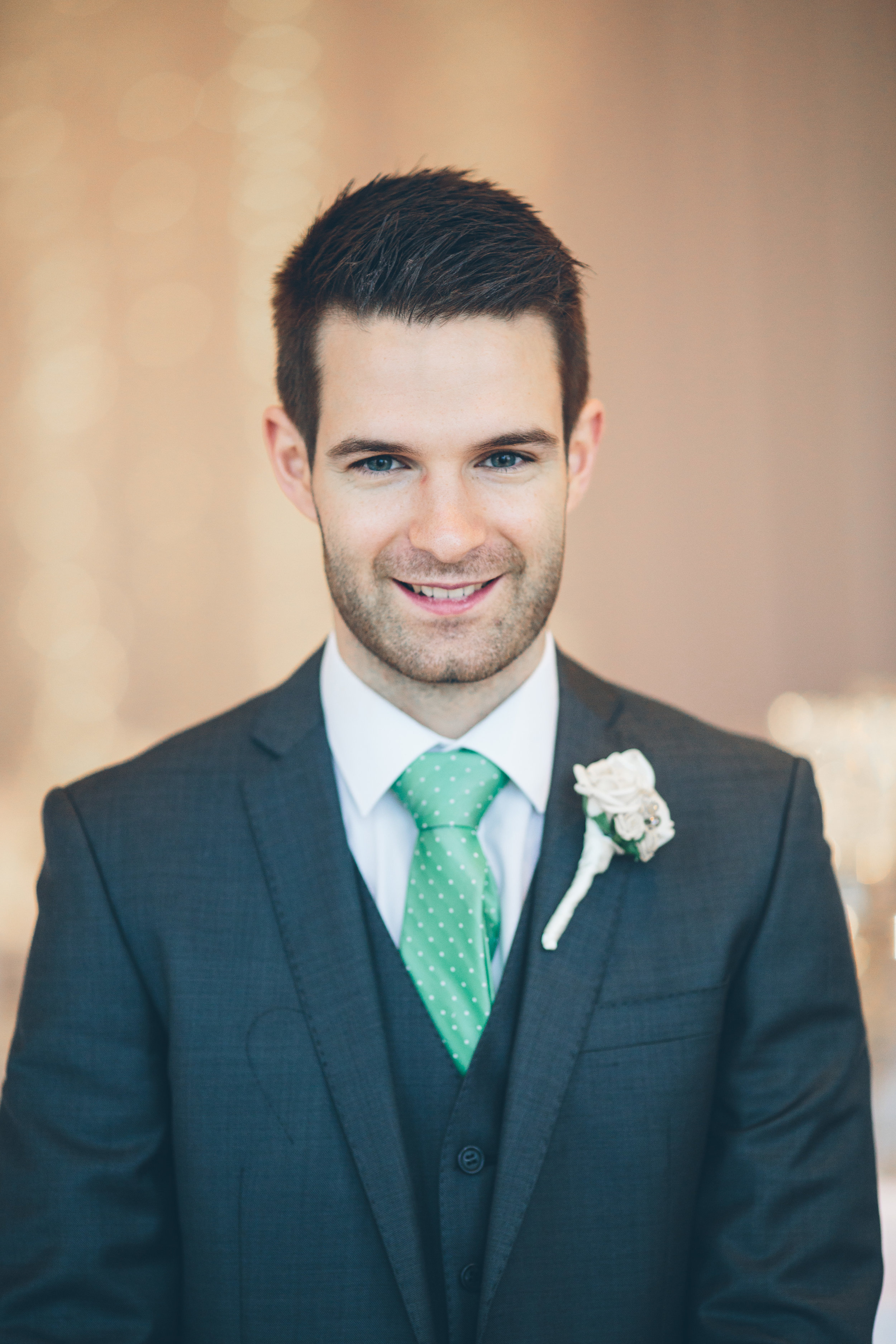 Portrait of a smiling man in a dark suit with a white shirt and a green polka-dot tie, wearing a white boutonniere on his lapel.