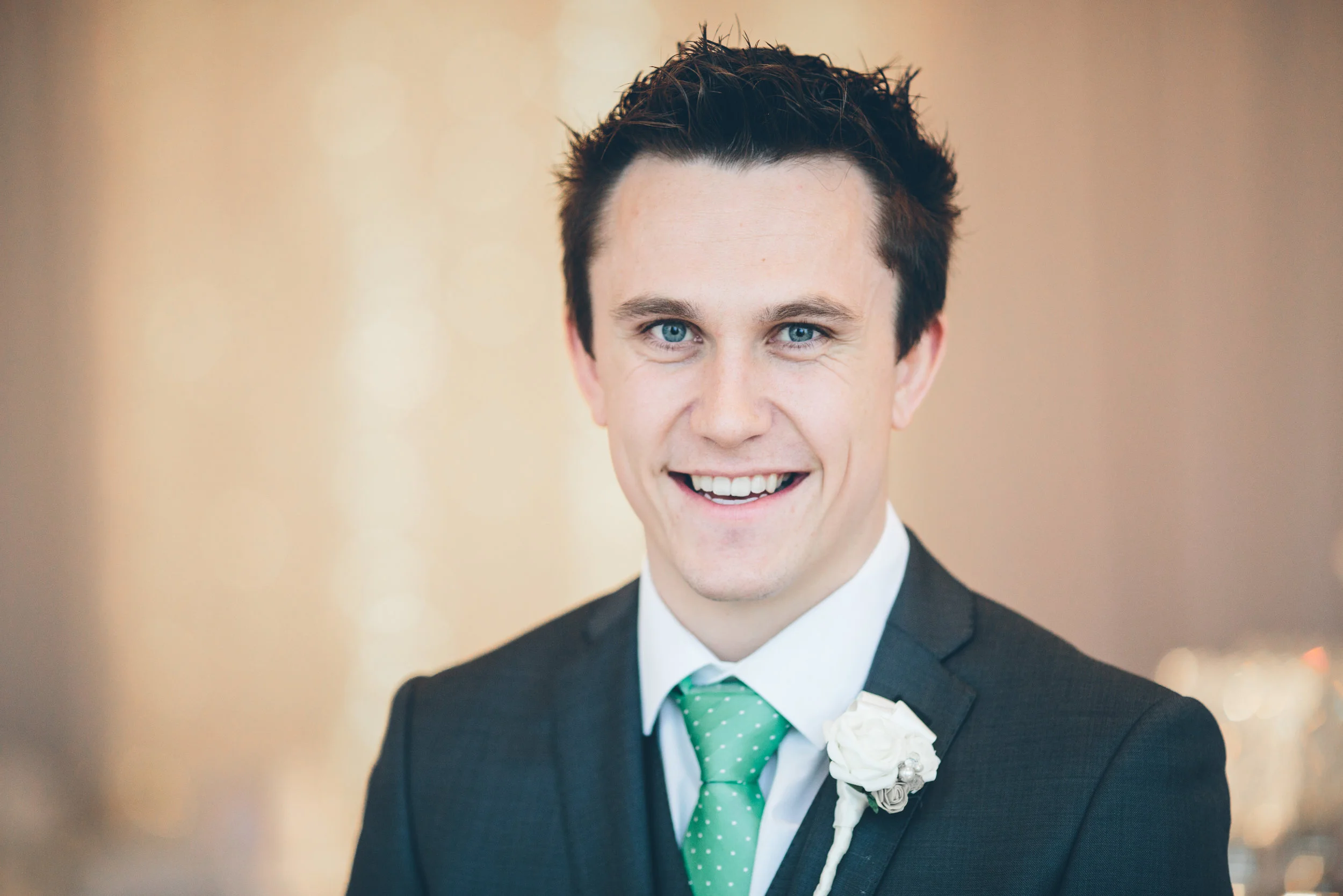 A young man with dark hair and blue eyes wearing a black suit, white shirt, and green polka dot tie, smiling at the camera. He has a white flower boutonniere on his lapel.