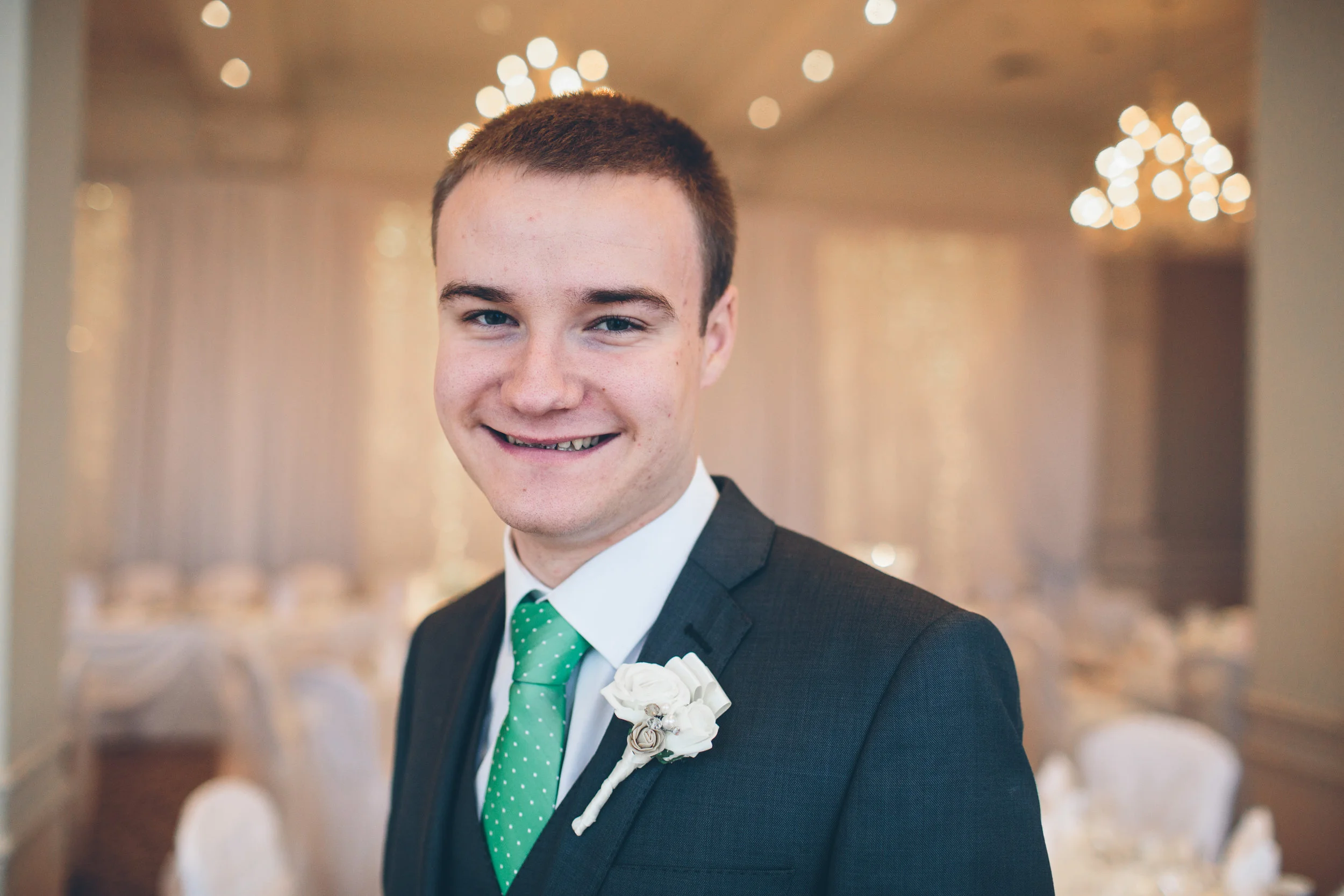 Young man in a suit with a green polka dot tie, smiling at the camera with chandeliers and decorated tables in the background.