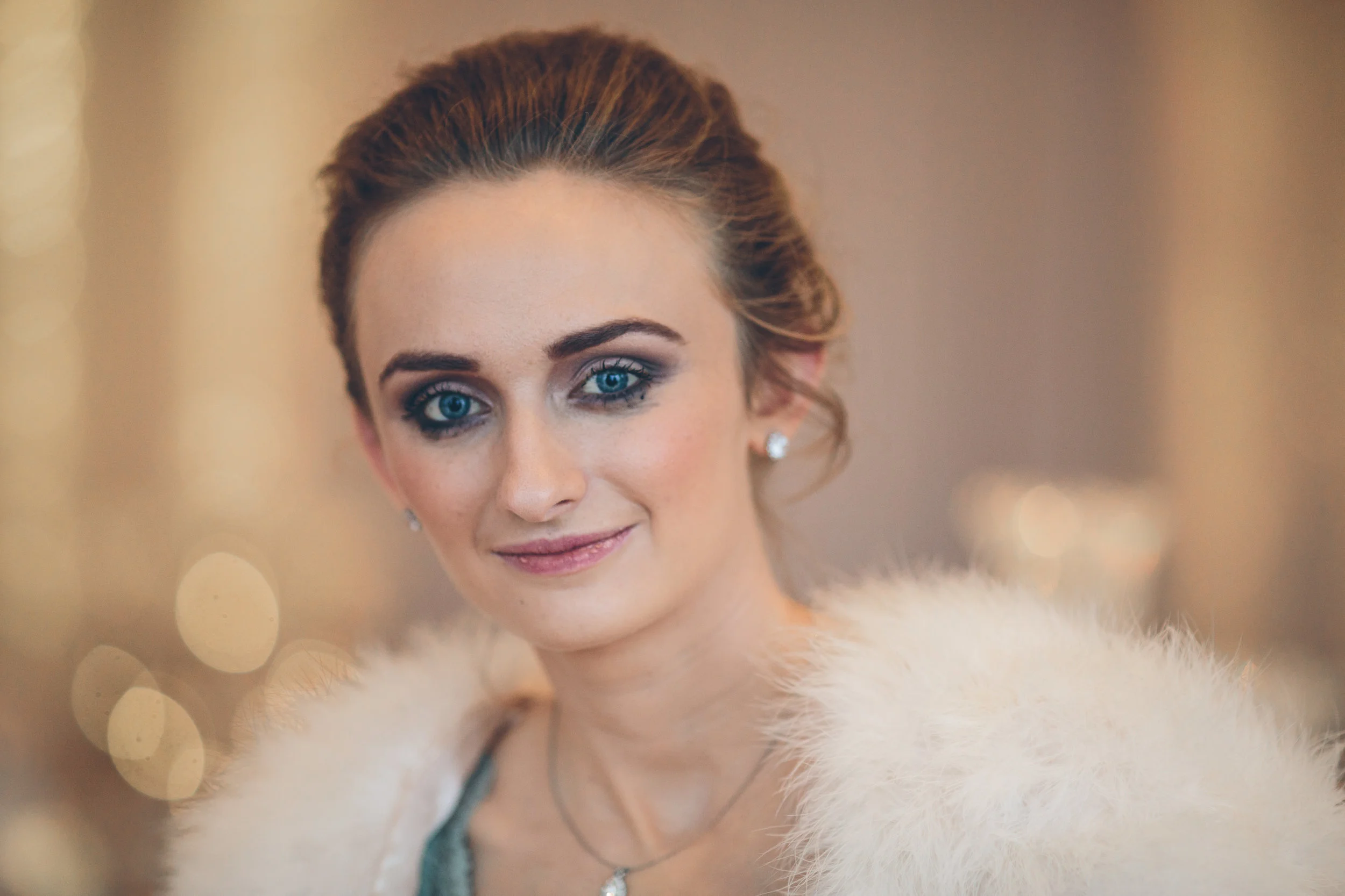 A woman with blue eyes and brown hair styled in an updo, wearing pearl earrings and a fur shawl, smiling.