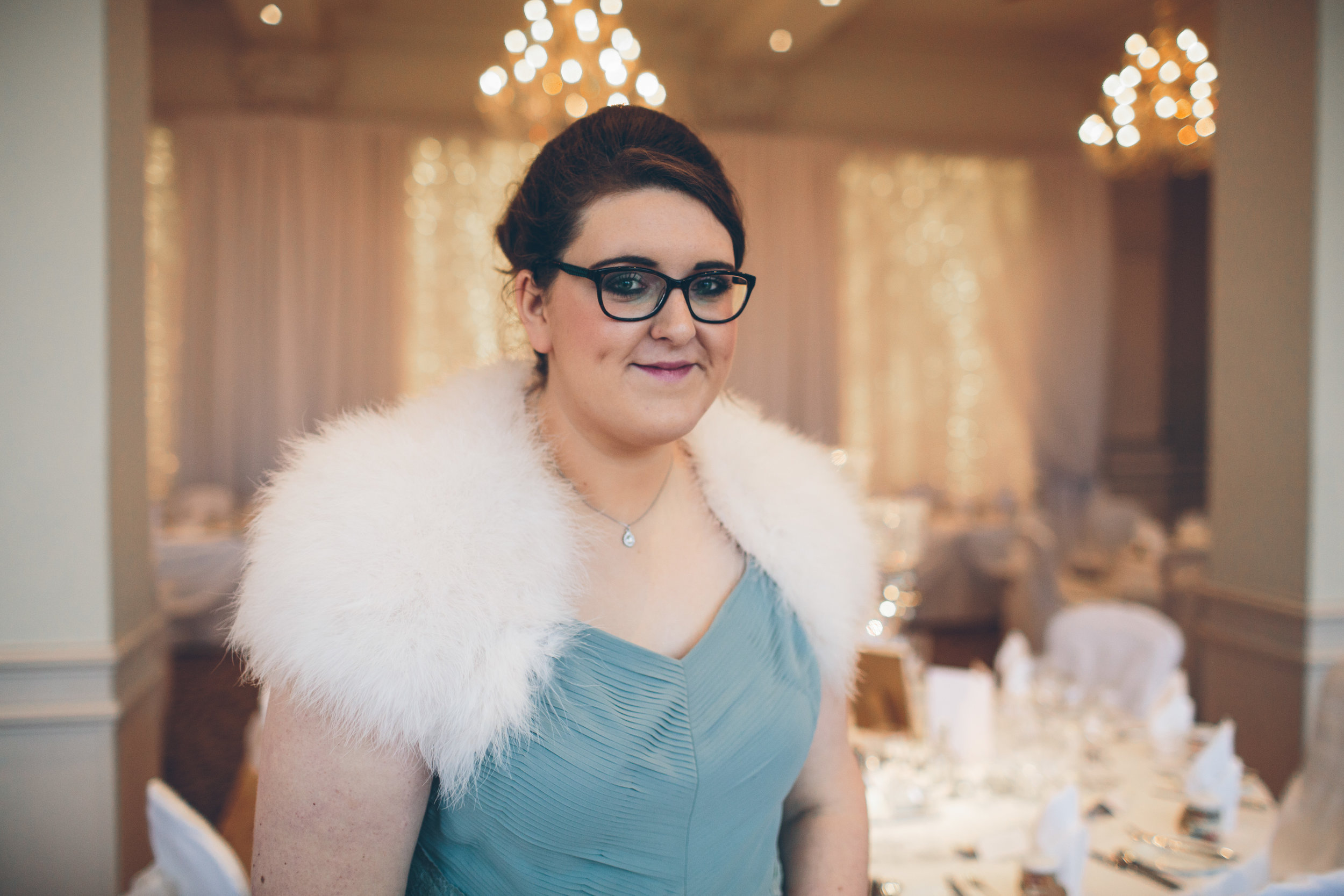 A woman with dark hair, glasses, and a white fur stole over a light blue dress, standing in a decorated banquet room with chandeliers and table settings.