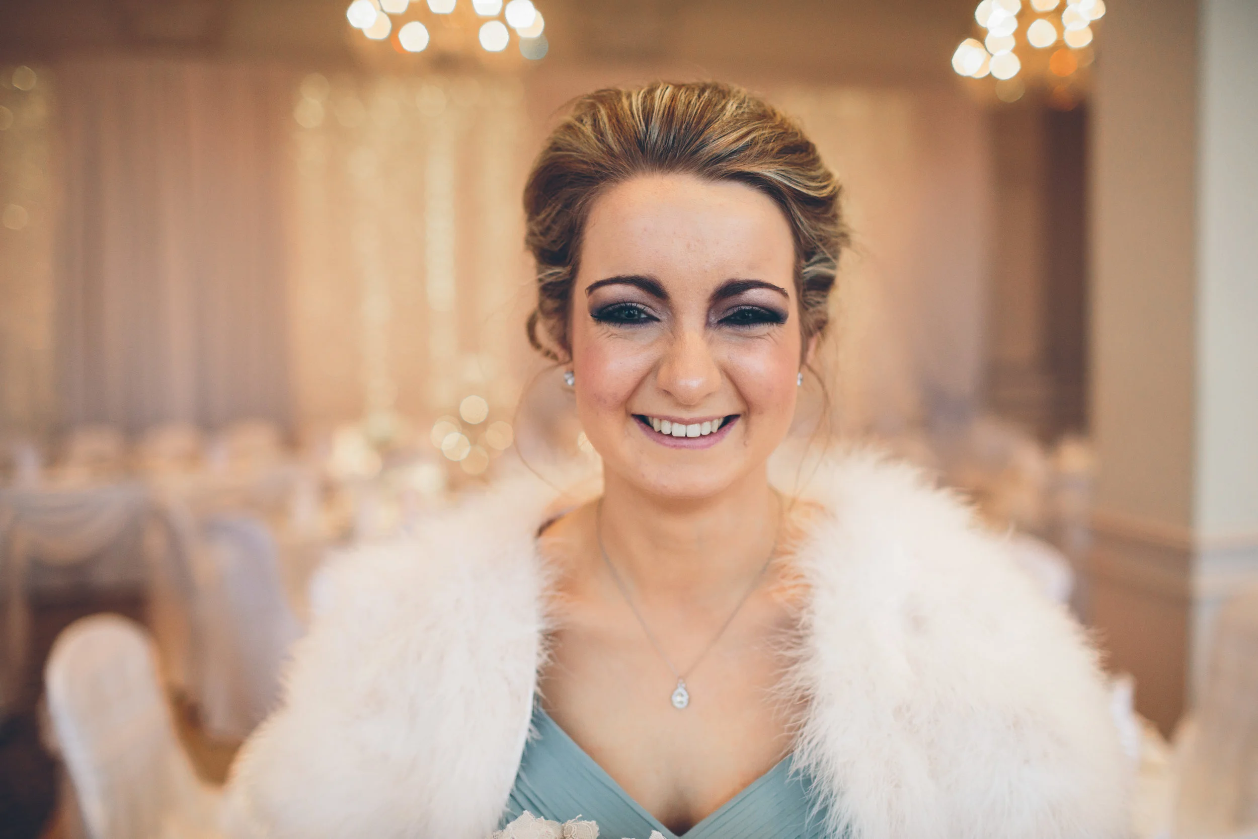 A smiling woman with makeup and styled hair, wearing a white fur stole and a light blue dress, standing in a decorated banquet hall with warm lighting and blurred background
