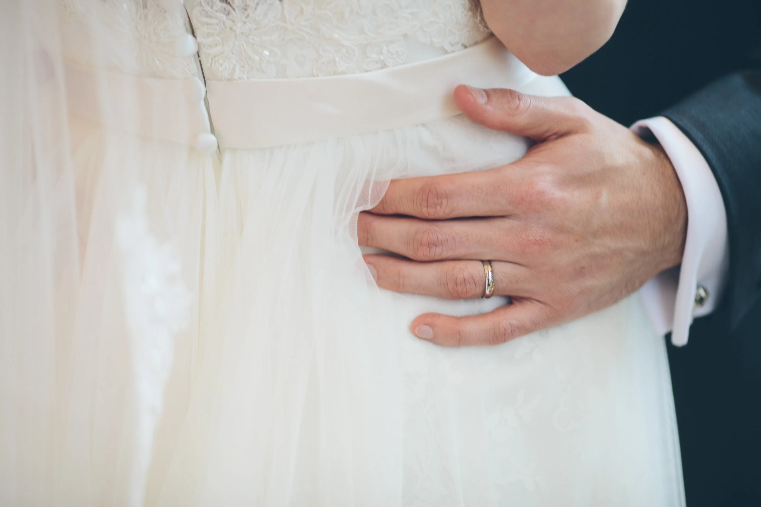 A groom gently holds a bride's waist during their wedding, with the bride wearing a white wedding gown and a wedding ring visible on the groom's finger.