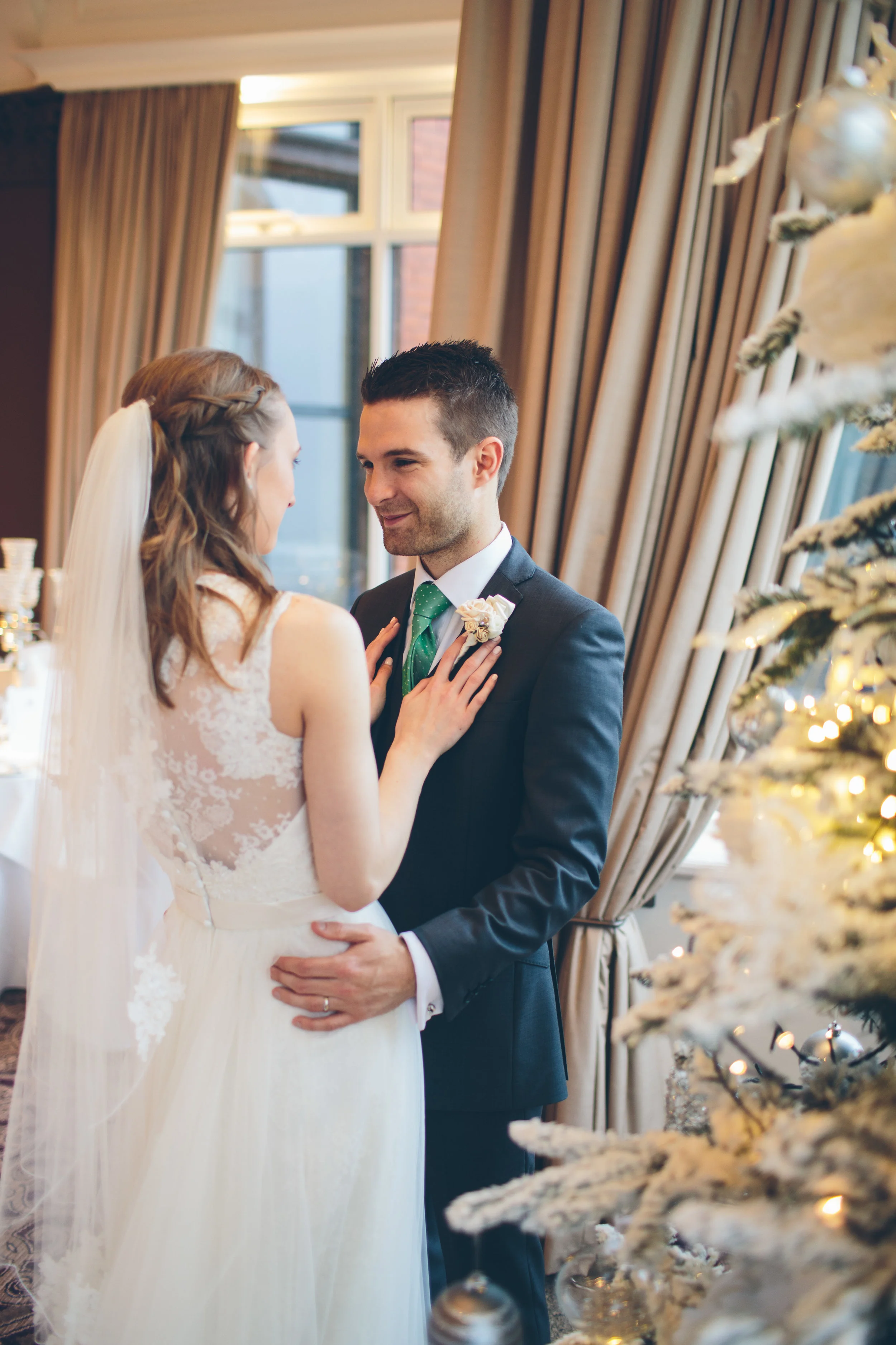 A bride and groom share a tender moment during their wedding, standing indoors near a decorated Christmas tree.