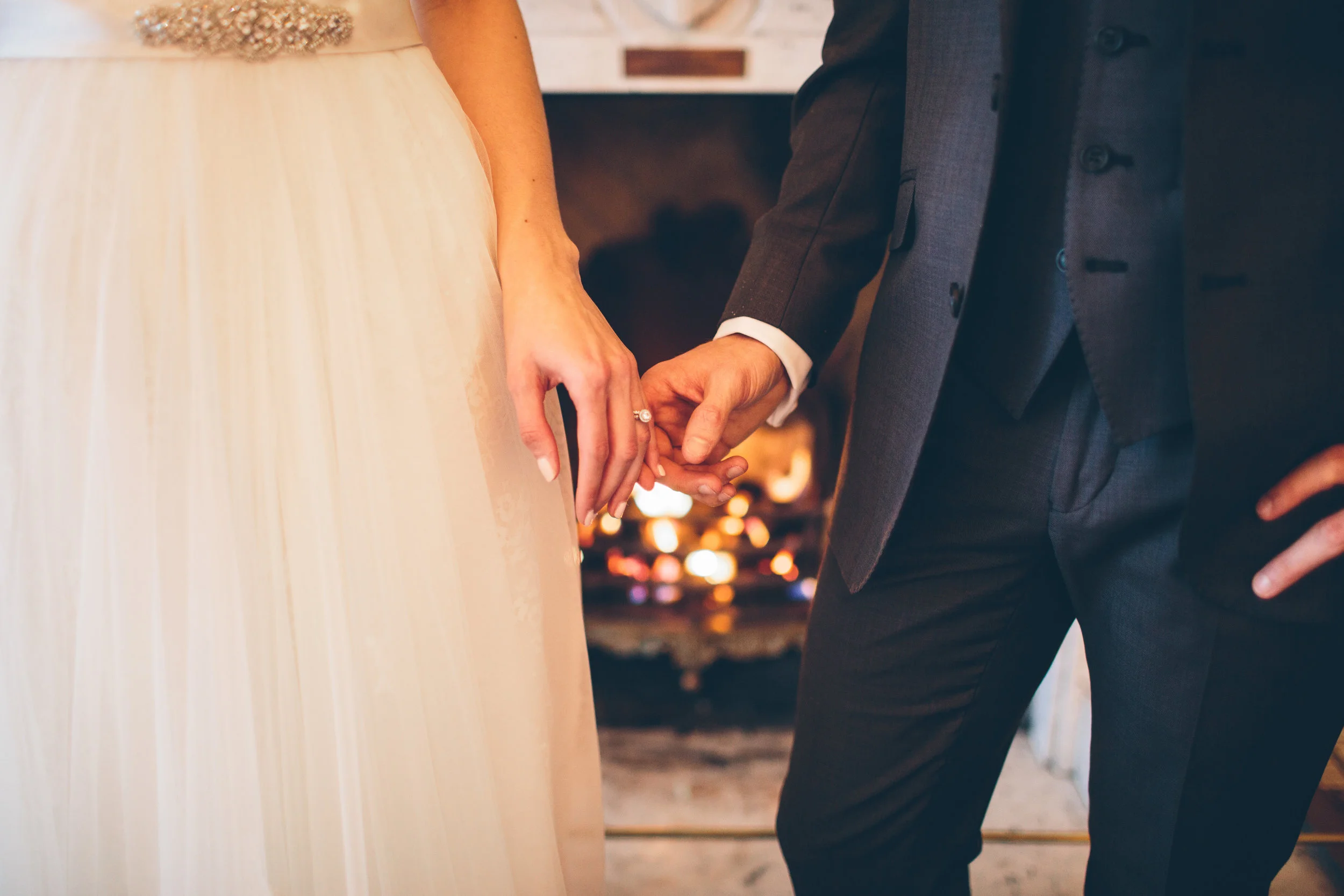 Close-up of a bride and groom holding hands during a wedding ceremony, with the bride's hand displaying a wedding ring and the groom wearing a gray suit, in front of a fireplace with lit candles.