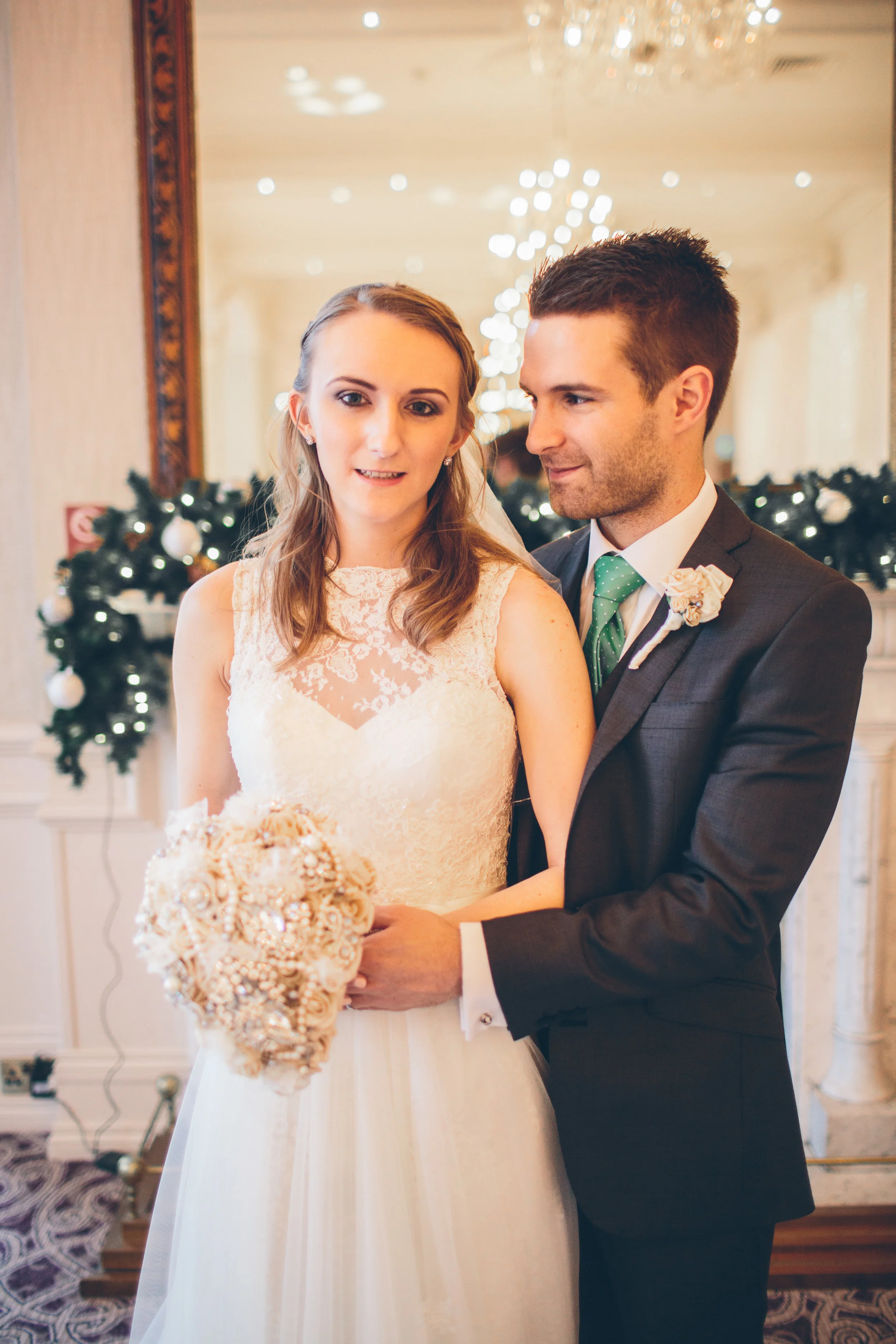 Bride and groom standing together, the bride holding a bouquet of flowers, in a decorated wedding venue with lights and garlands in the background.