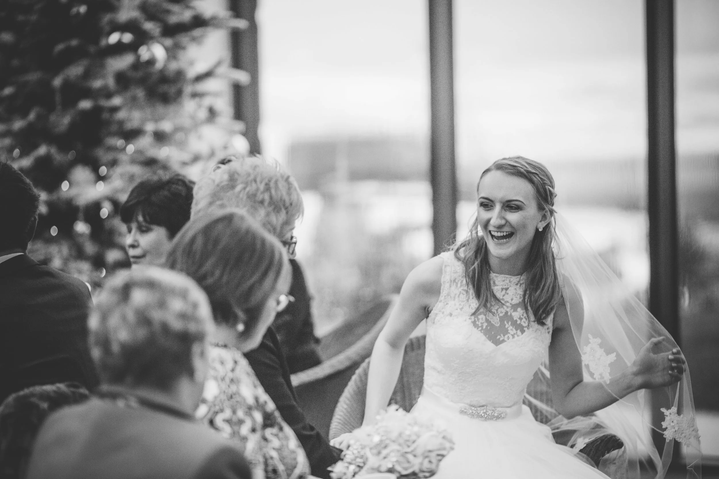 A smiling bride in a wedding dress sitting with a group of people at a wedding reception, with a decorated Christmas tree in the background.