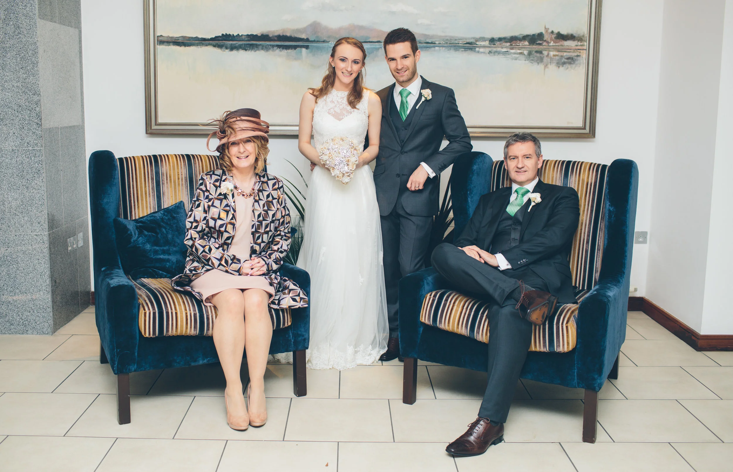 A wedding photo with five people. A bride and groom stand in the center, with a woman sitting on the left and a man sitting on the right. The woman on the left is wearing a patterned jacket, a beige dress, and a fancy hat. The man on the right is dre