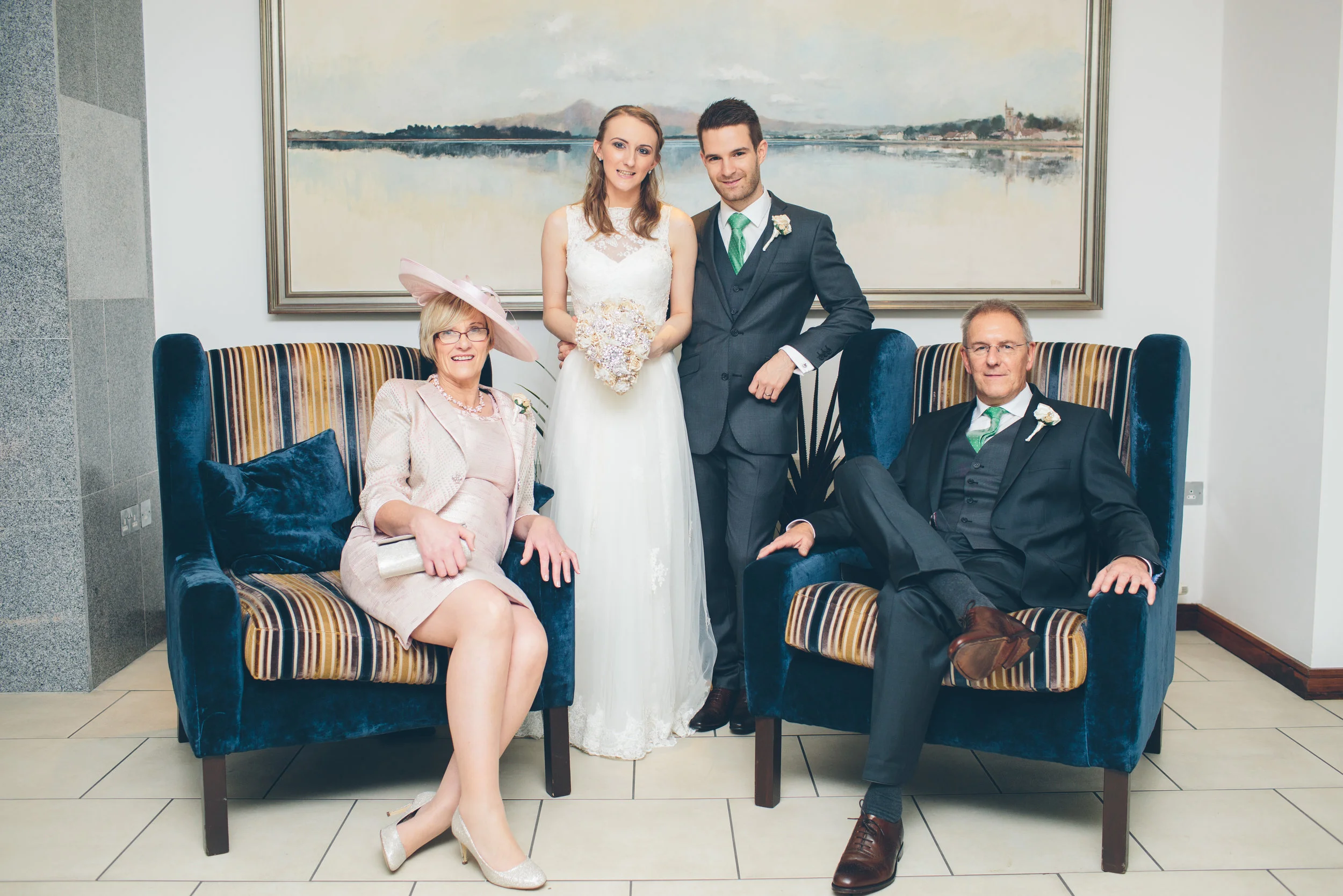 A wedding party with a bride and groom standing behind seated older woman and man, all dressed in formal attire, in an indoor setting with a large landscape painting on the wall behind them.