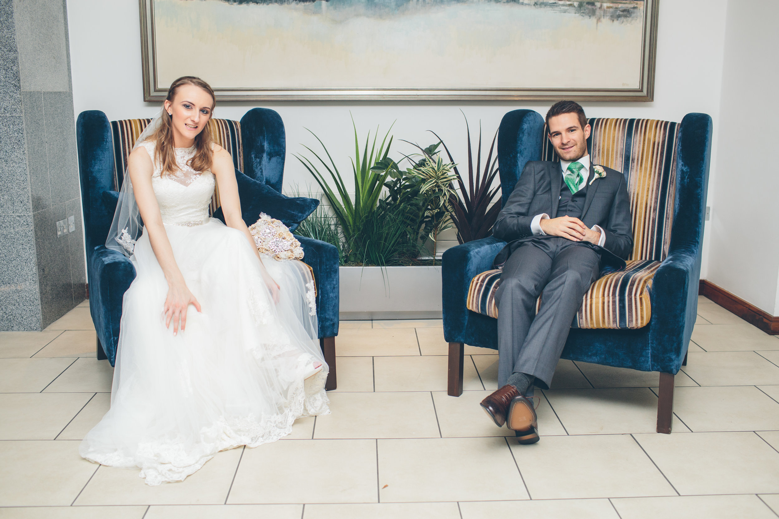 Bride and groom sitting on blue velvet chairs indoors, with plants and a large painting behind them.