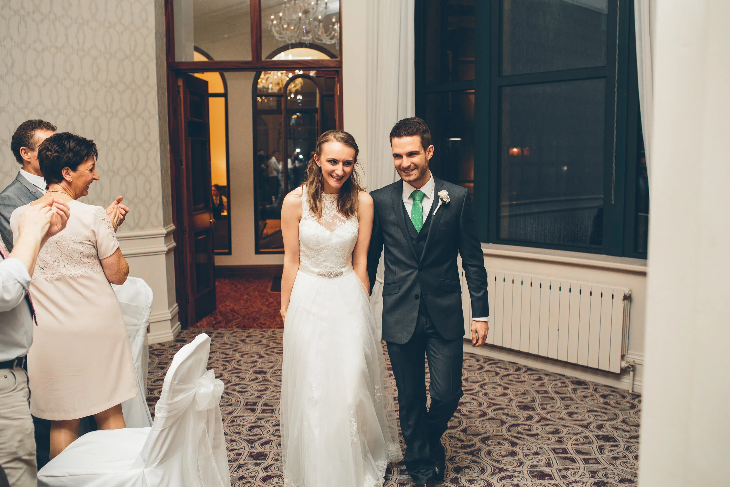 A bride and groom walking together in a reception hall, smiling. The bride wears a white wedding dress and the groom wears a dark suit with a green tie. Guests are applauding and celebrating in the background.