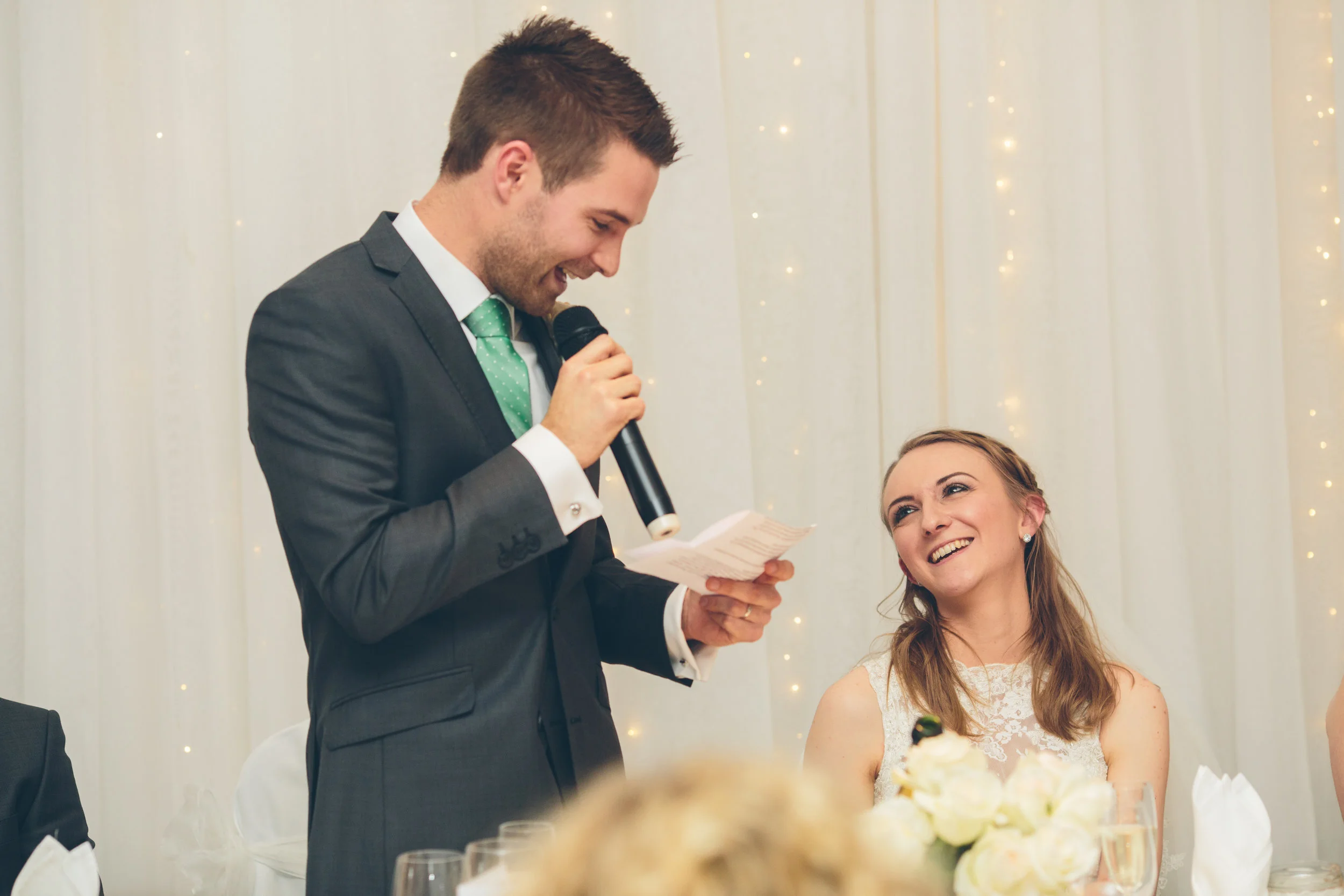 A man in a suit giving a speech at a wedding reception, smiling at a woman in a white dress who is sitting at the table and smiling back.