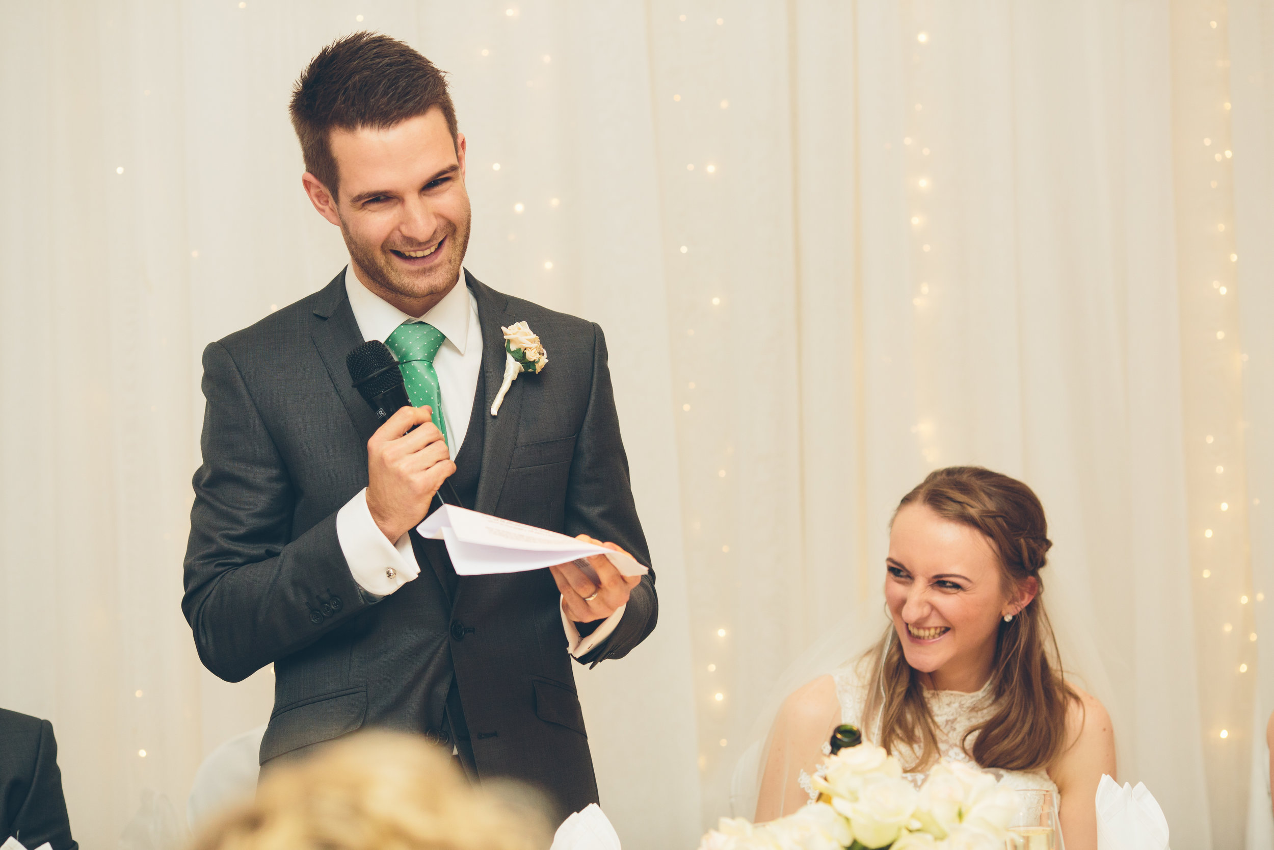 A man in a gray suit with a green tie giving a speech at a wedding reception, holding a microphone and notes, while the bride, sitting and smiling, listens with a bouquet in front of her.
