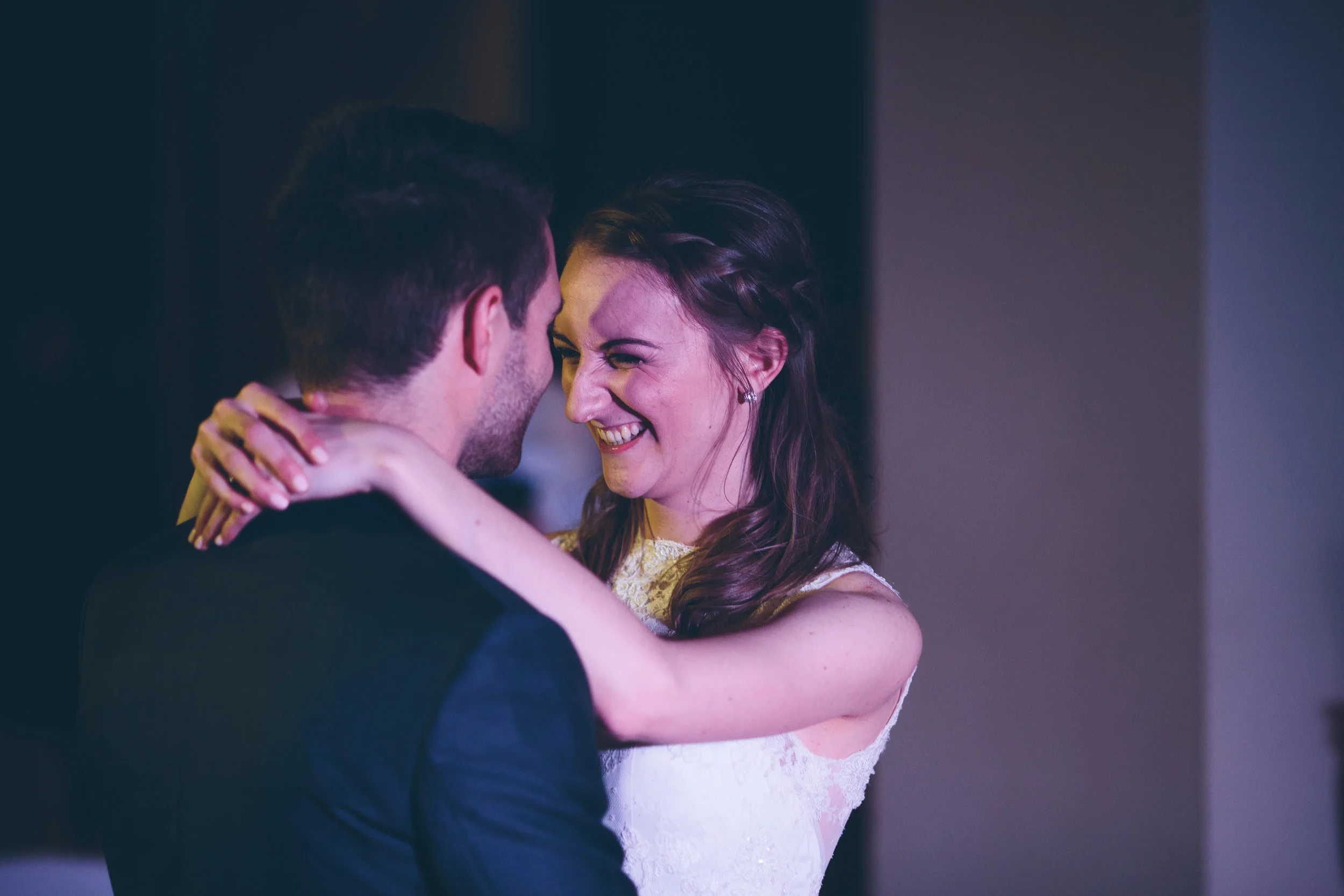 A couple dancing and smiling at each other, with the woman wearing a white dress with lace details and the man in a dark suit, in a dimly lit setting.