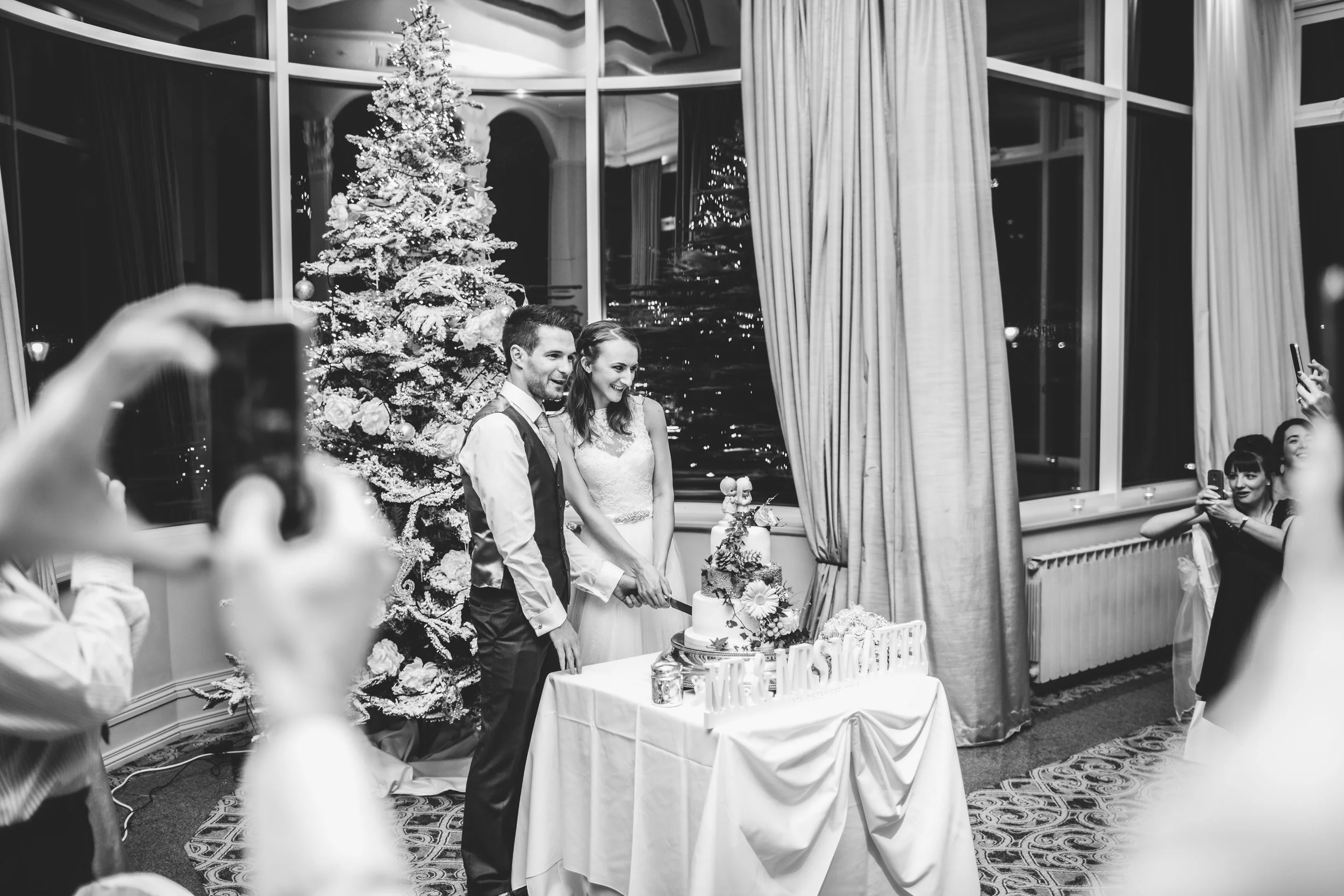 A bride and groom cutting a wedding cake at their celebration, with a decorated Christmas tree and large windows in the background, surrounded by guests taking photos.