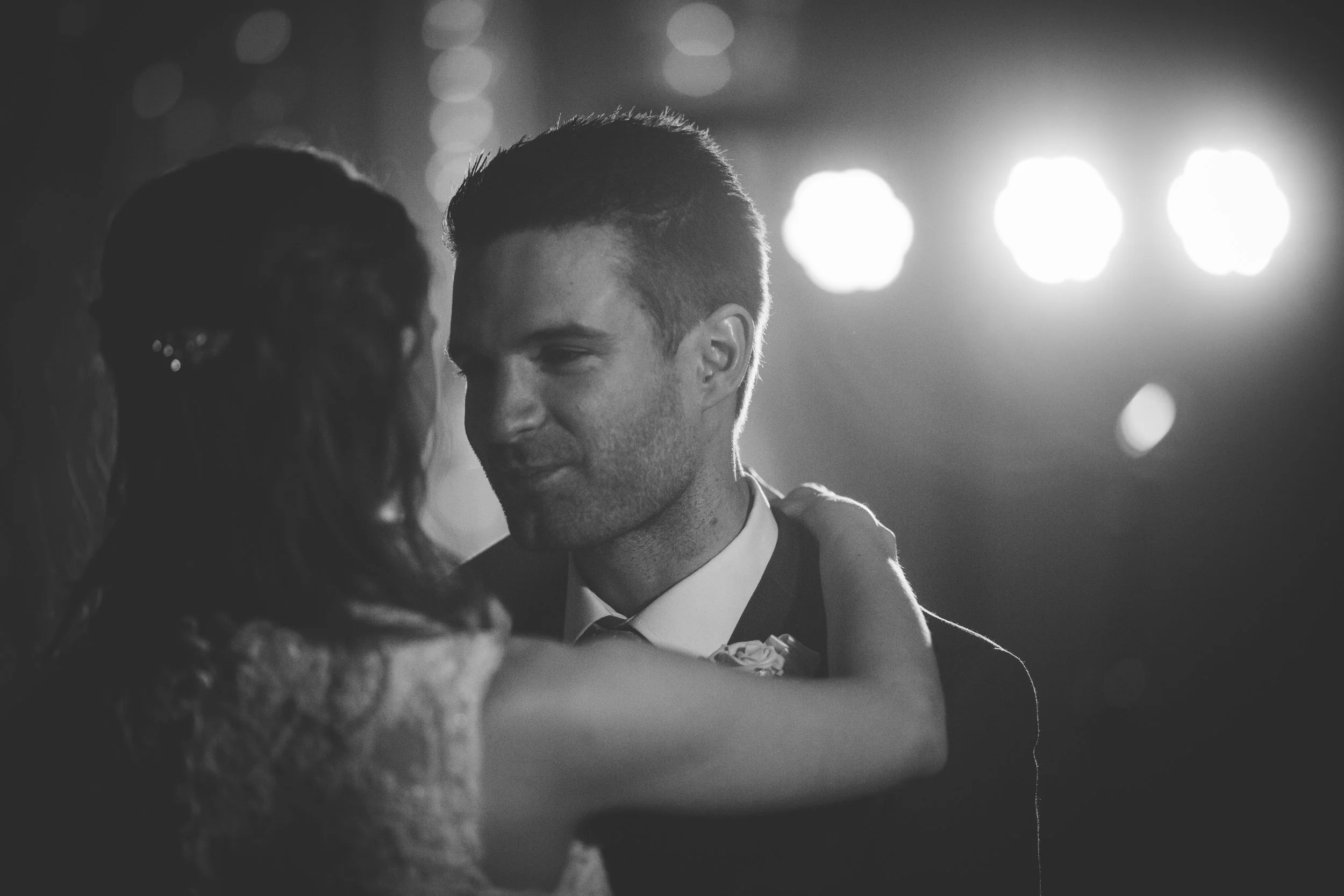 A black-and-white photo of a couple dancing closely with gentle smiles, illuminated by soft lights.