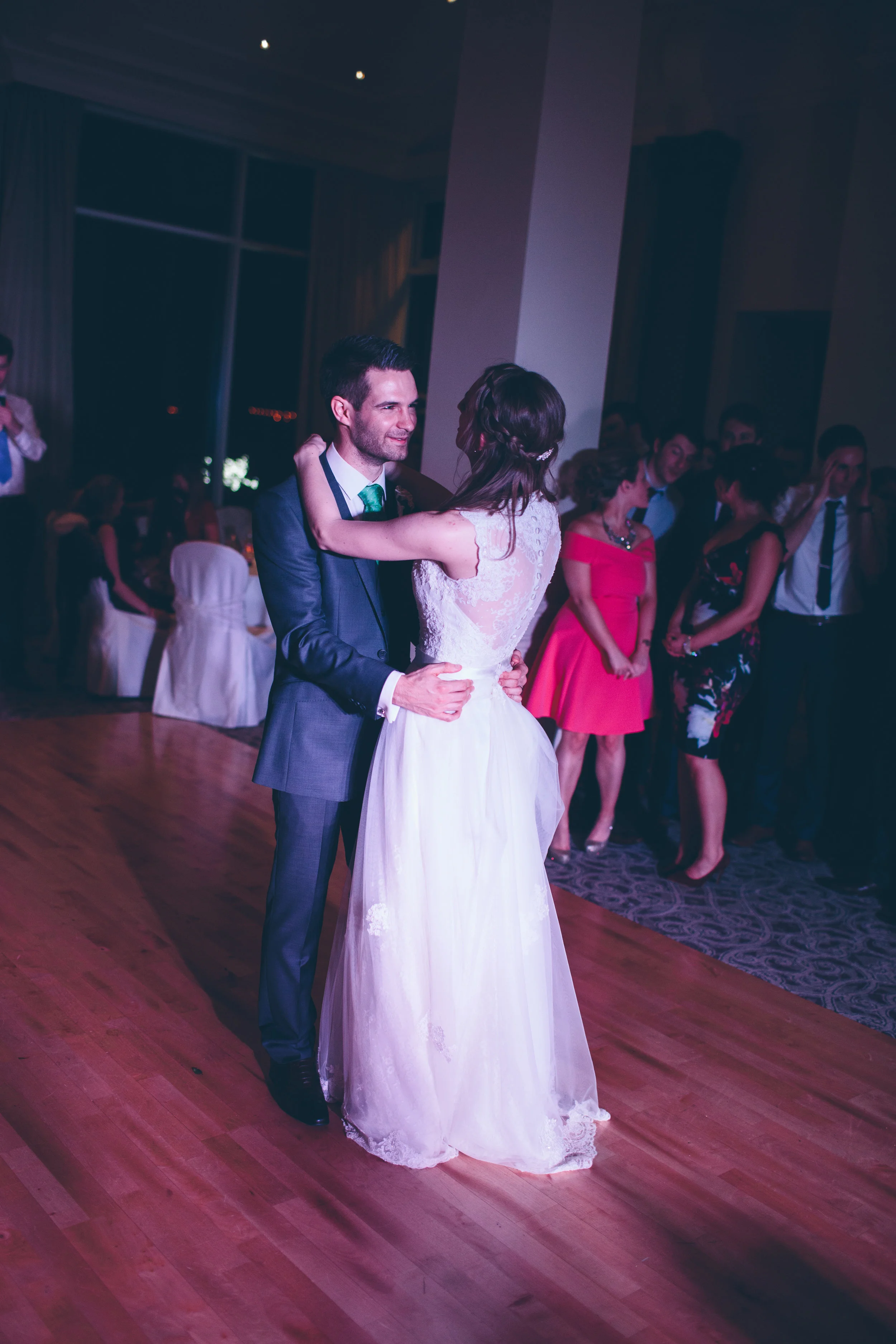A bride and groom are dancing at their wedding reception, surrounded by friends and family in a dimly lit ballroom.
