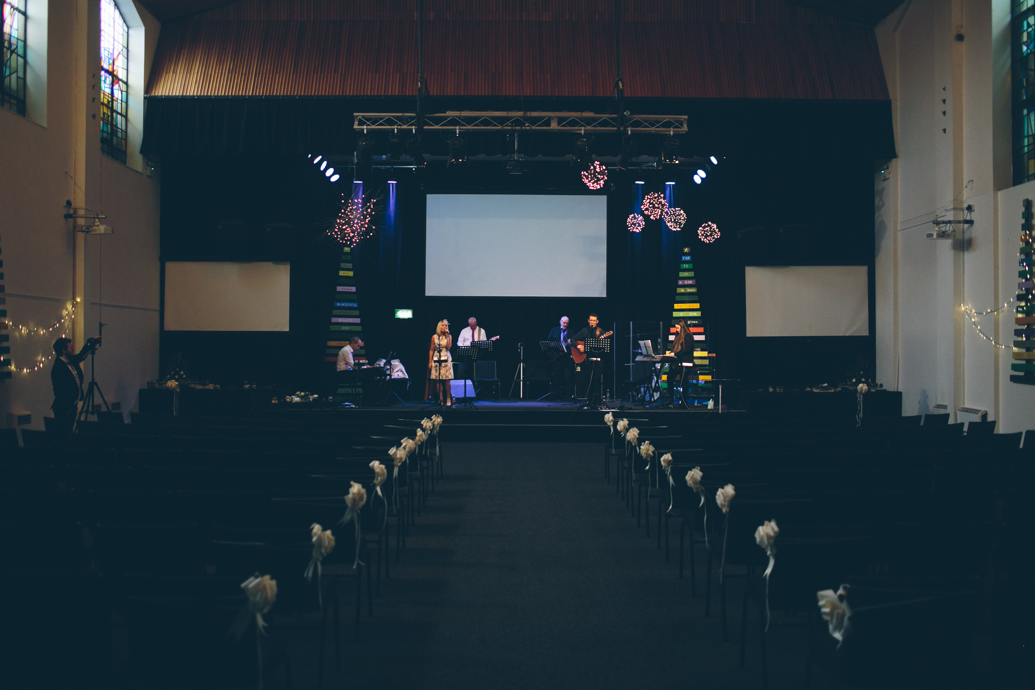 Stage with a band performing in an auditorium decorated for a celebration or event, with chairs decorated with bows in the foreground.