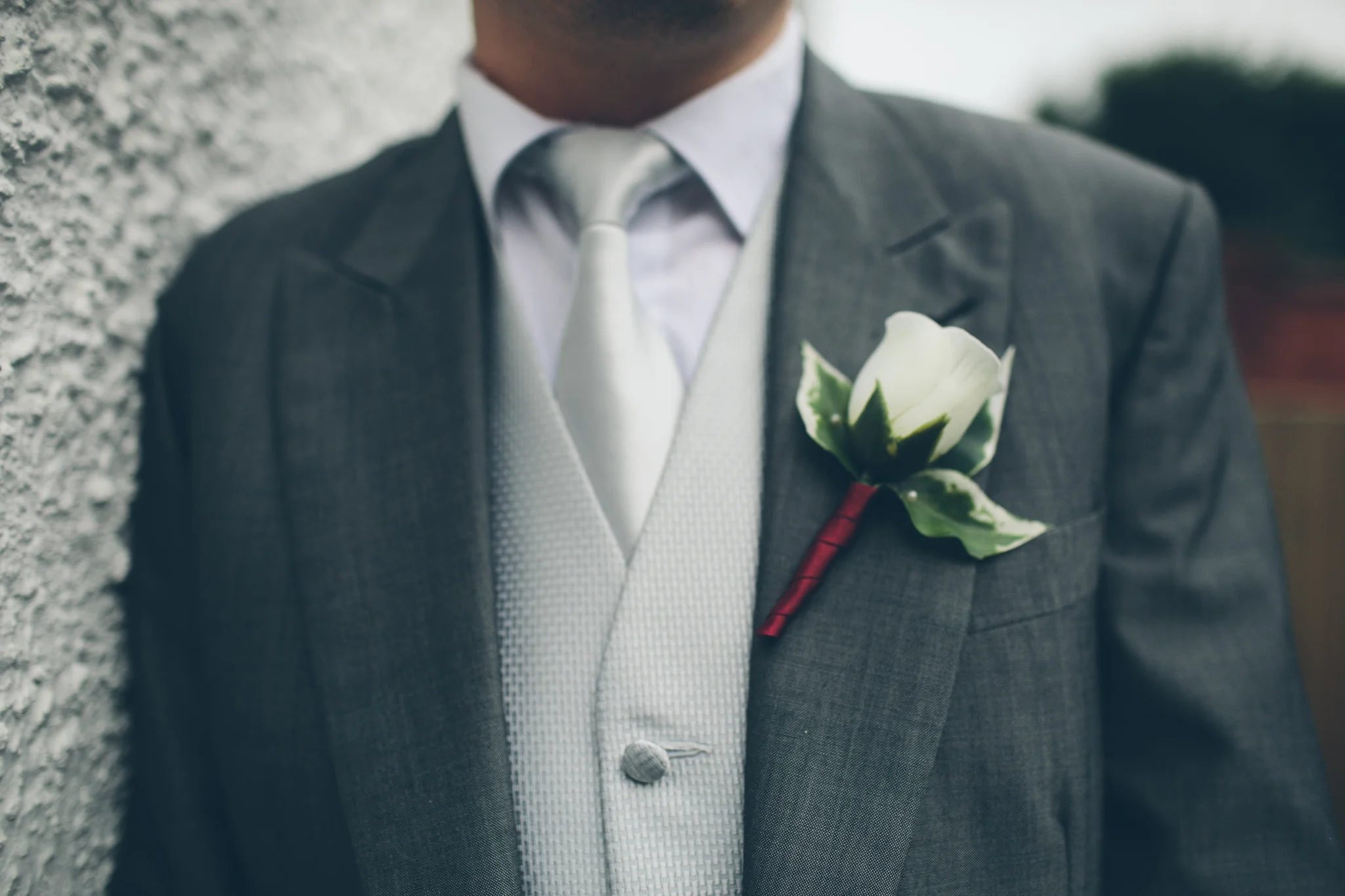 Close-up of a man wearing a gray suit with a white shirt and a white tie, with a white flower boutonniere on the left lapel.