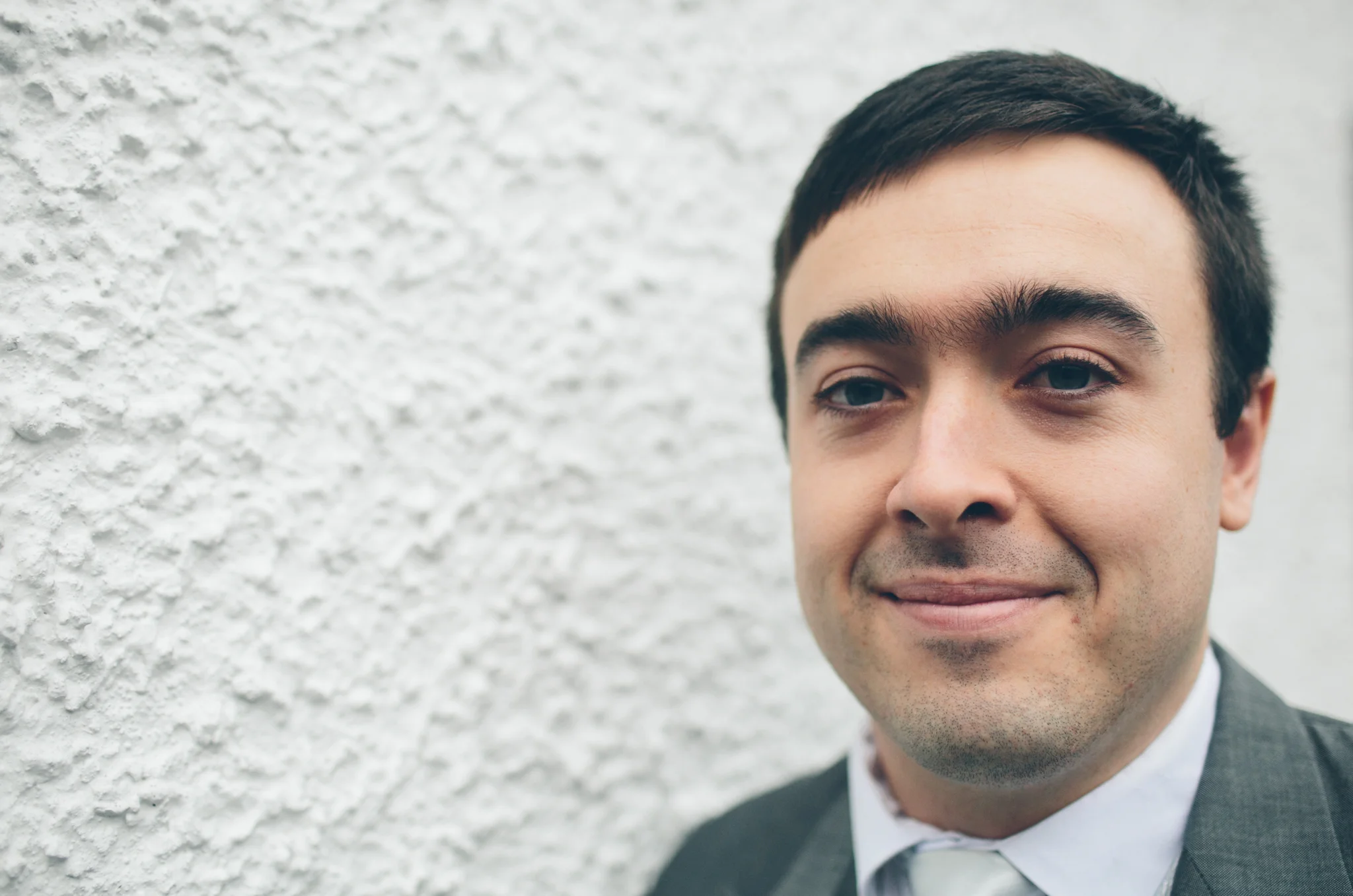 A close-up of a man with short dark hair, wearing a suit and tie, smiling slightly in front of a textured white wall.
