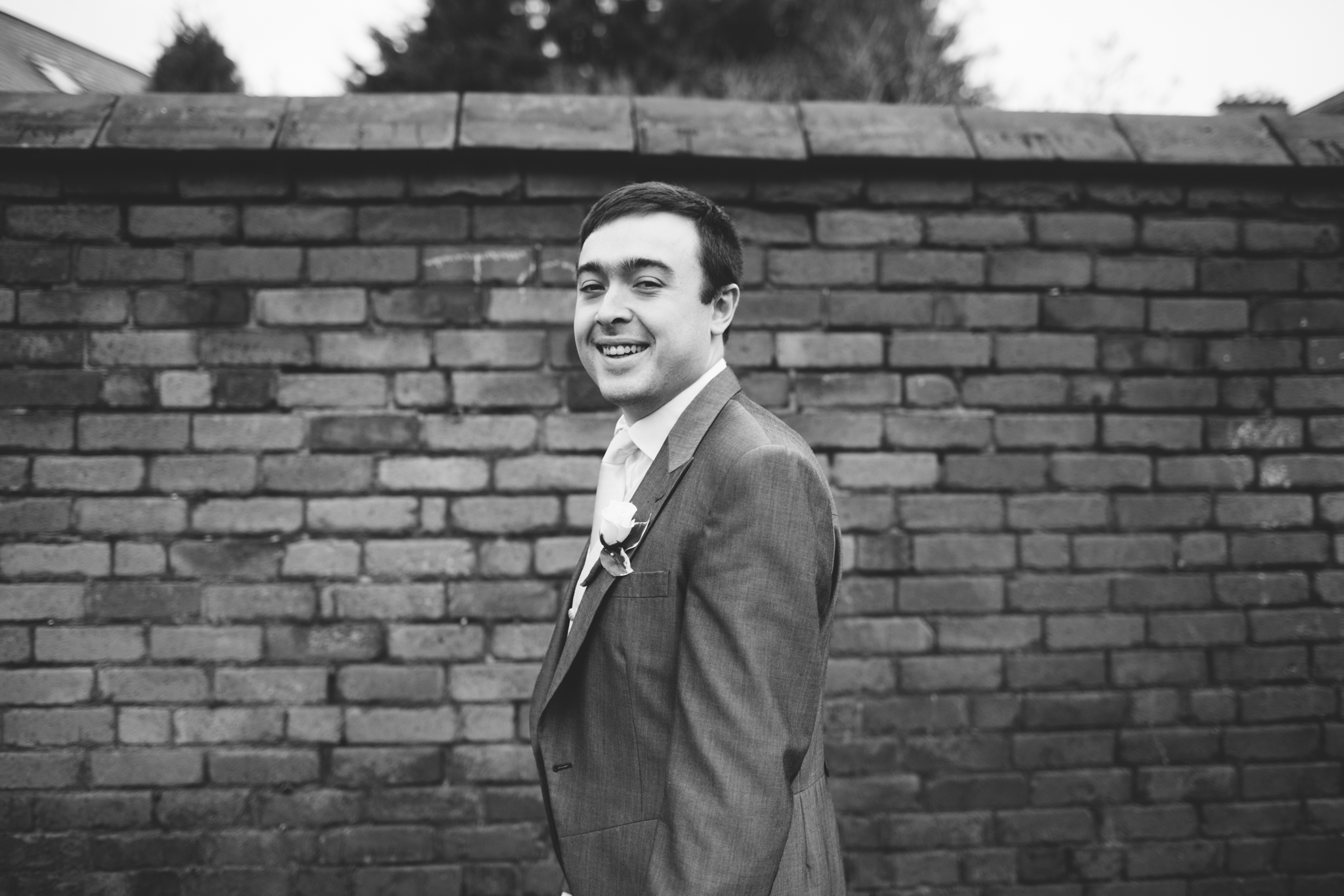 A young man dressed in a suit with a boutonniere, smiling and standing in front of a brick wall in black and white.