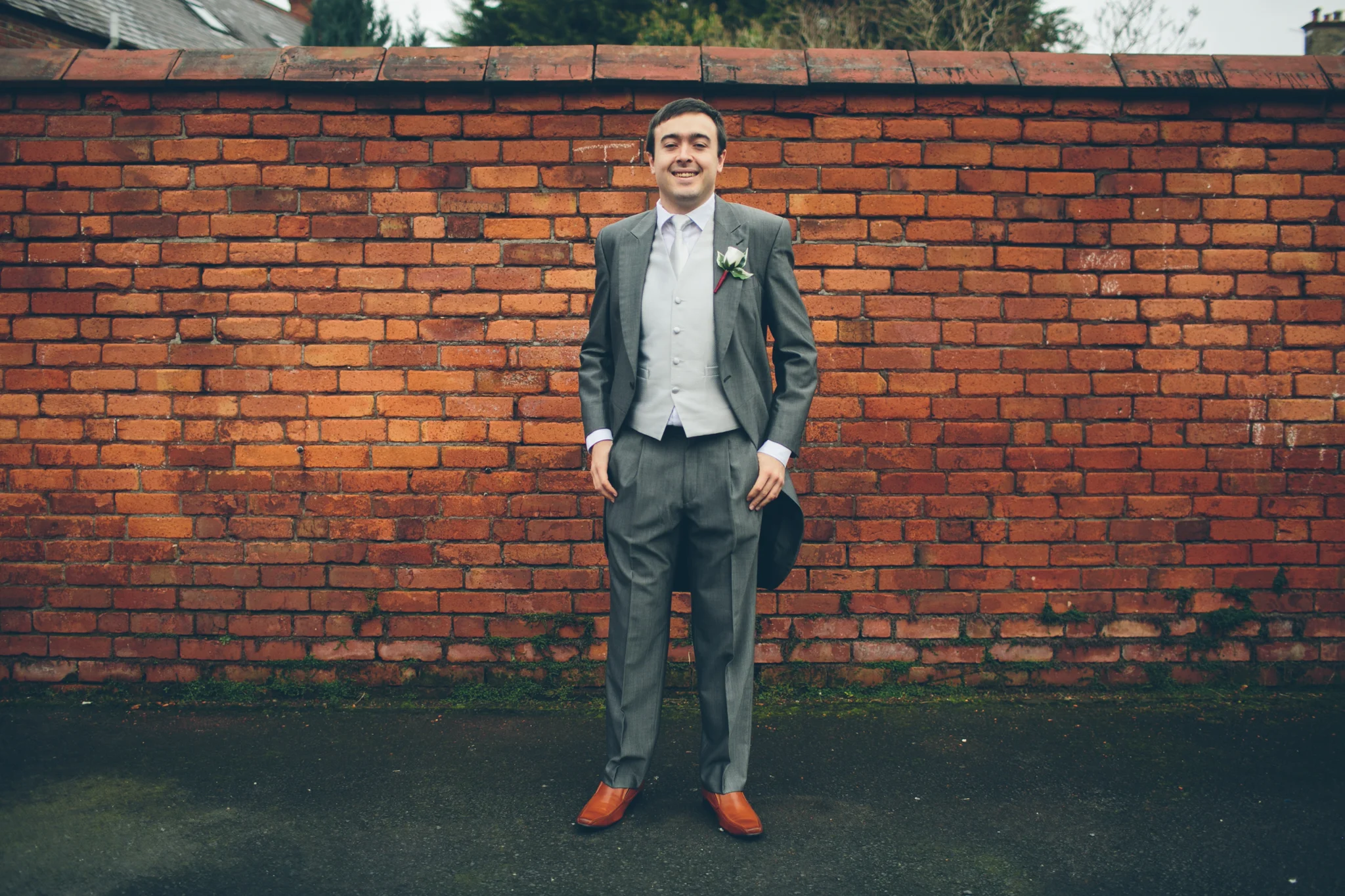 A man in a gray suit, white vest, and white shirt standing outdoors in front of a red brick wall. He has a boutonnière on his left lapel and is smiling. The ground appears wet.